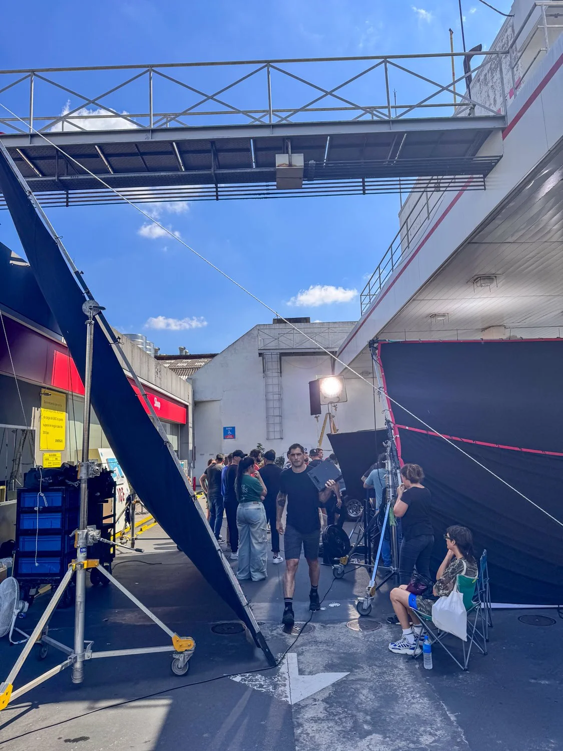 Behind-the-scenes view of a film or photo shoot with crew members and equipment, including lighting and black screens, outdoors under a blue sky.