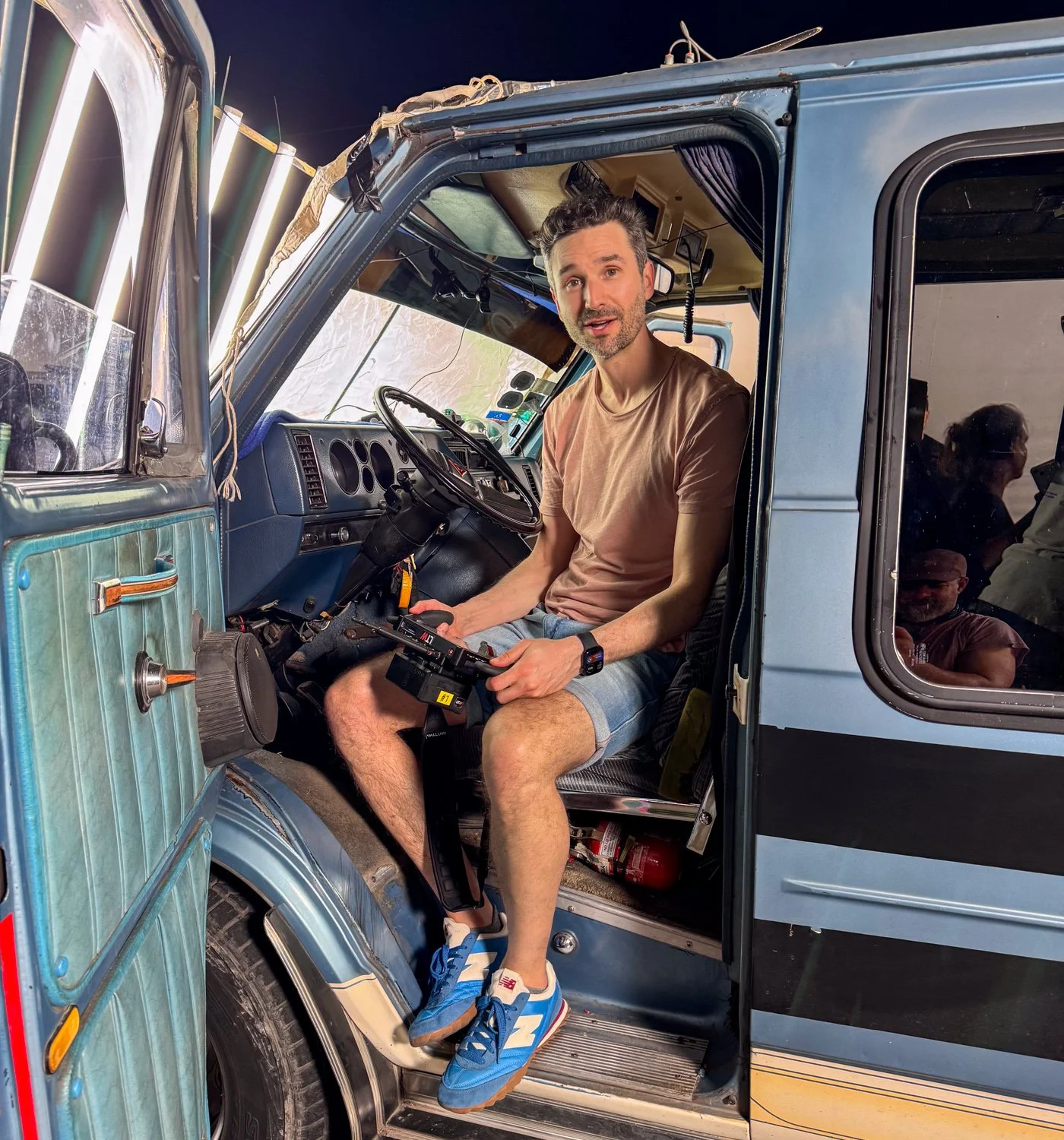 A man sitting inside the driver's seat of a blue utility vehicle, holding a remote control, with a surprised expression, wearing a beige T-shirt, shorts, and blue sneakers.
