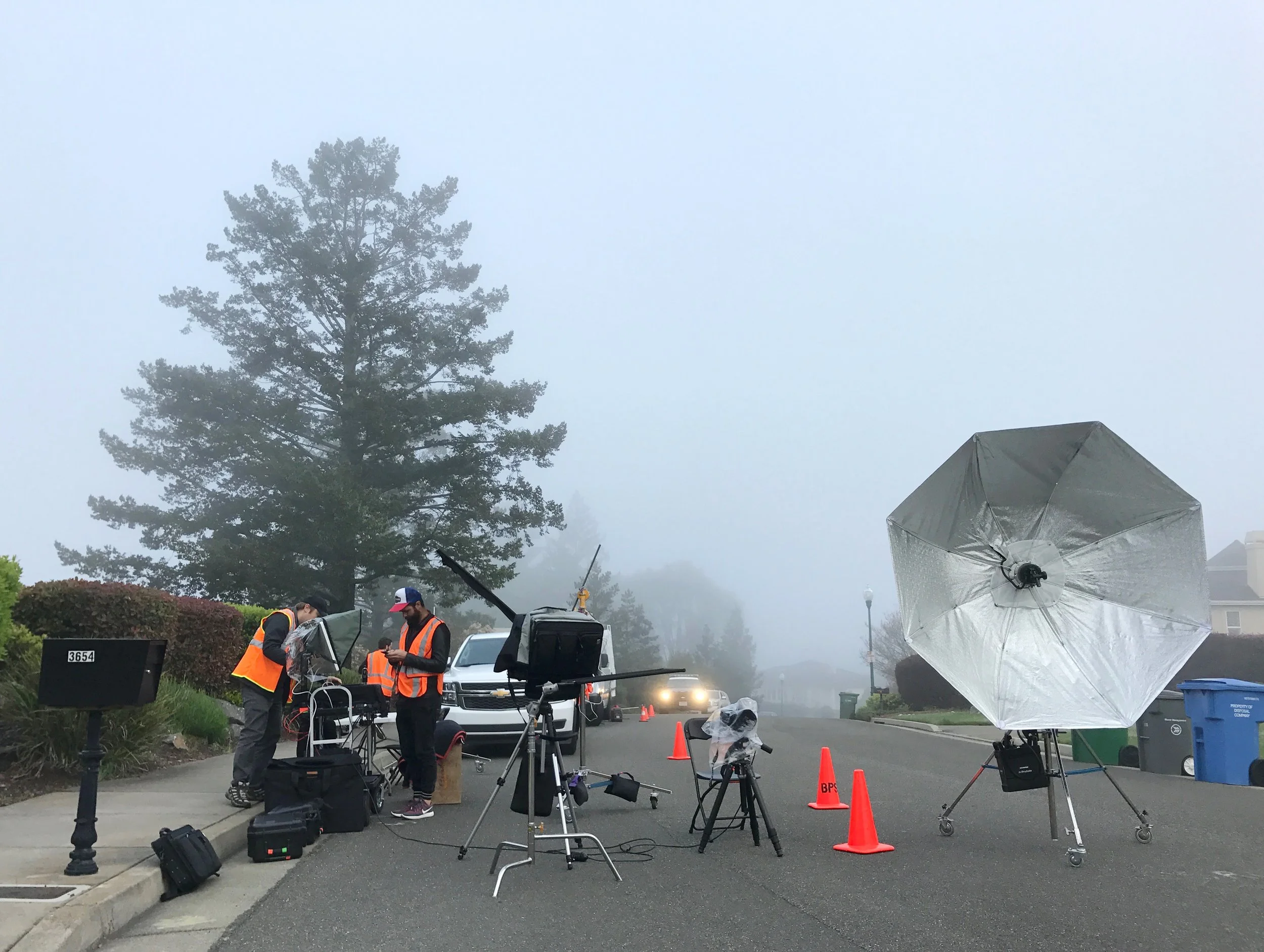 A photography setup on a foggy street with three people in orange vests working around cameras, lighting equipment, and umbrellas, with orange traffic cones and a blue recycling bin nearby.