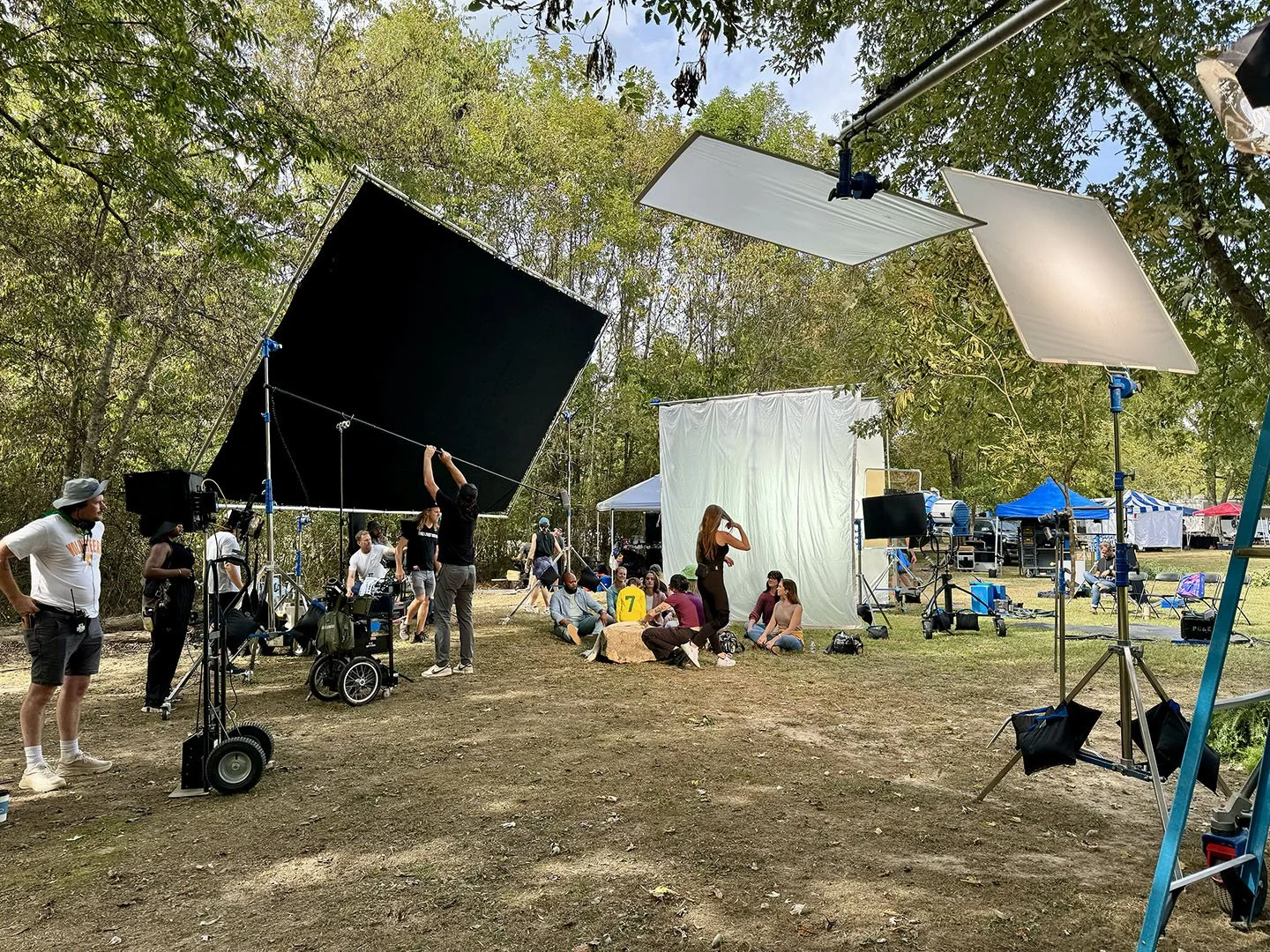 Film crew on outdoor set with lighting equipment, large black and white screens, and actors sitting on the ground, surrounded by trees and tents.