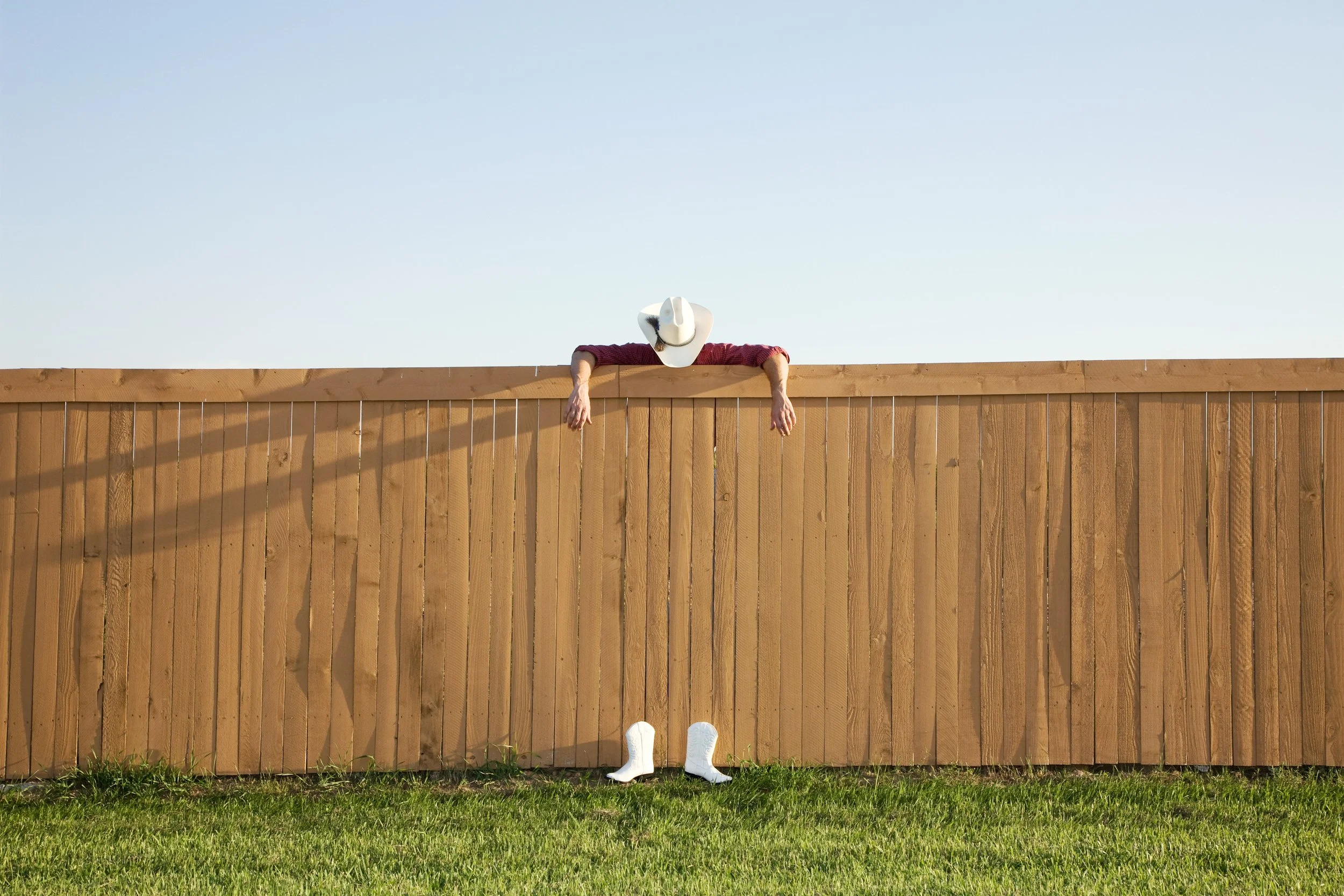A person wearing a cowboy hat leaning over a wooden fence with boots on the ground and arms hanging down, with a clear blue sky in the background.