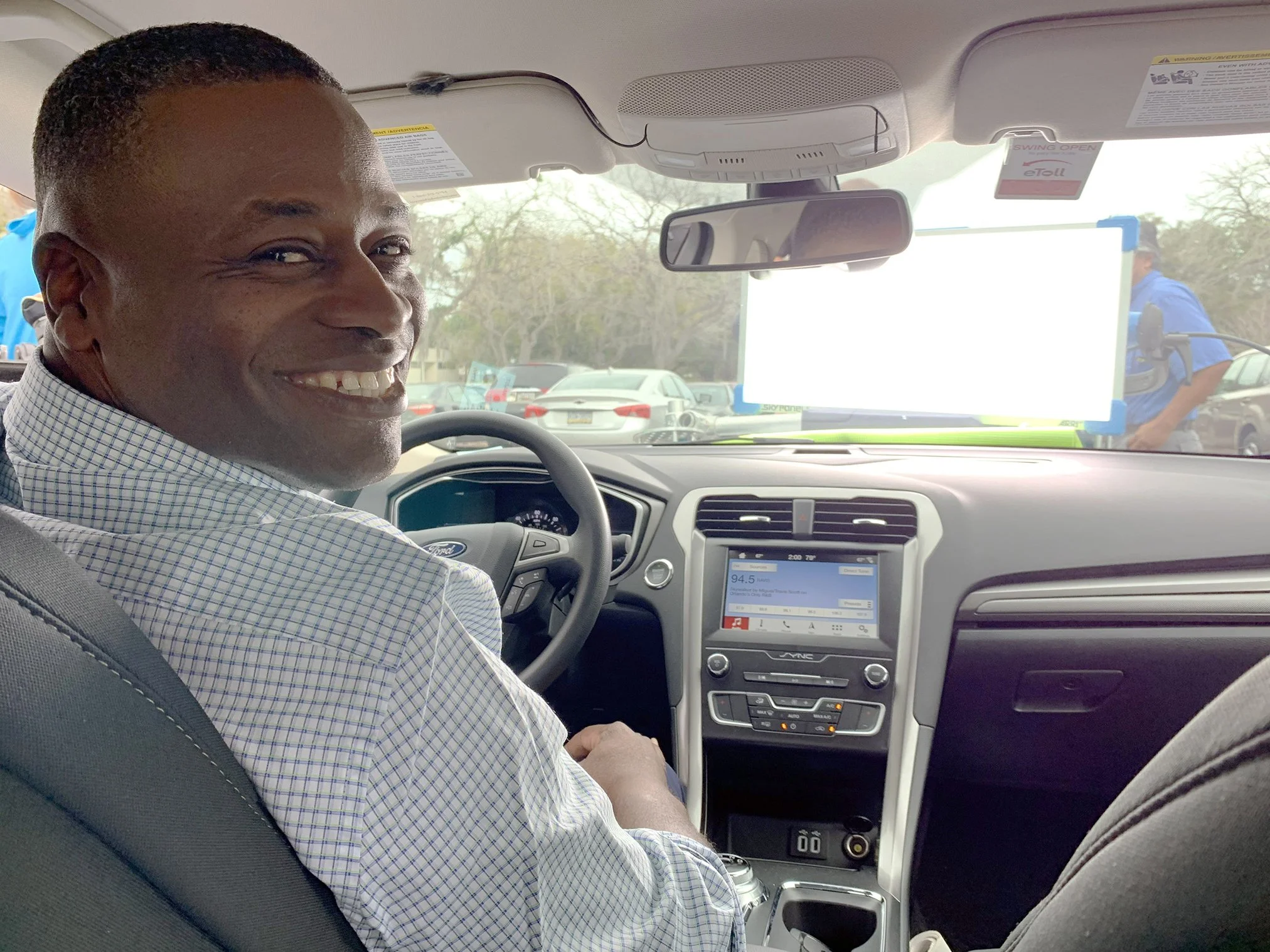 A man is smiling and sitting in the driver's seat of a vehicle, looking back over his shoulder, with a parking lot visible through the windshield.