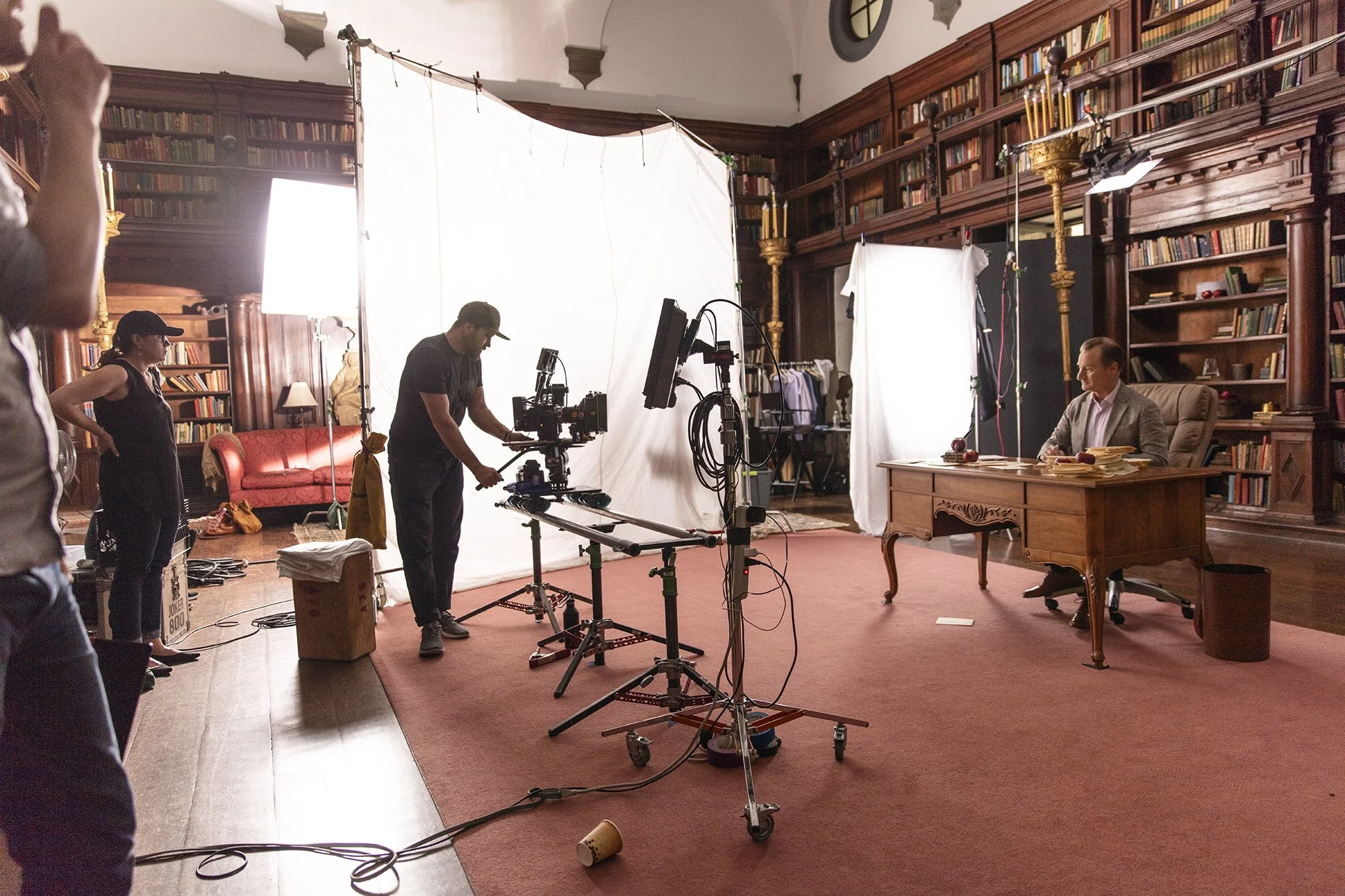 A film set in a large library with wooden bookshelves. A man in a gray suit is sitting at a wooden desk with stacks of papers, lighting equipment and white backdrop screens surrounding him. Crew members are adjusting camera and lighting equipment.