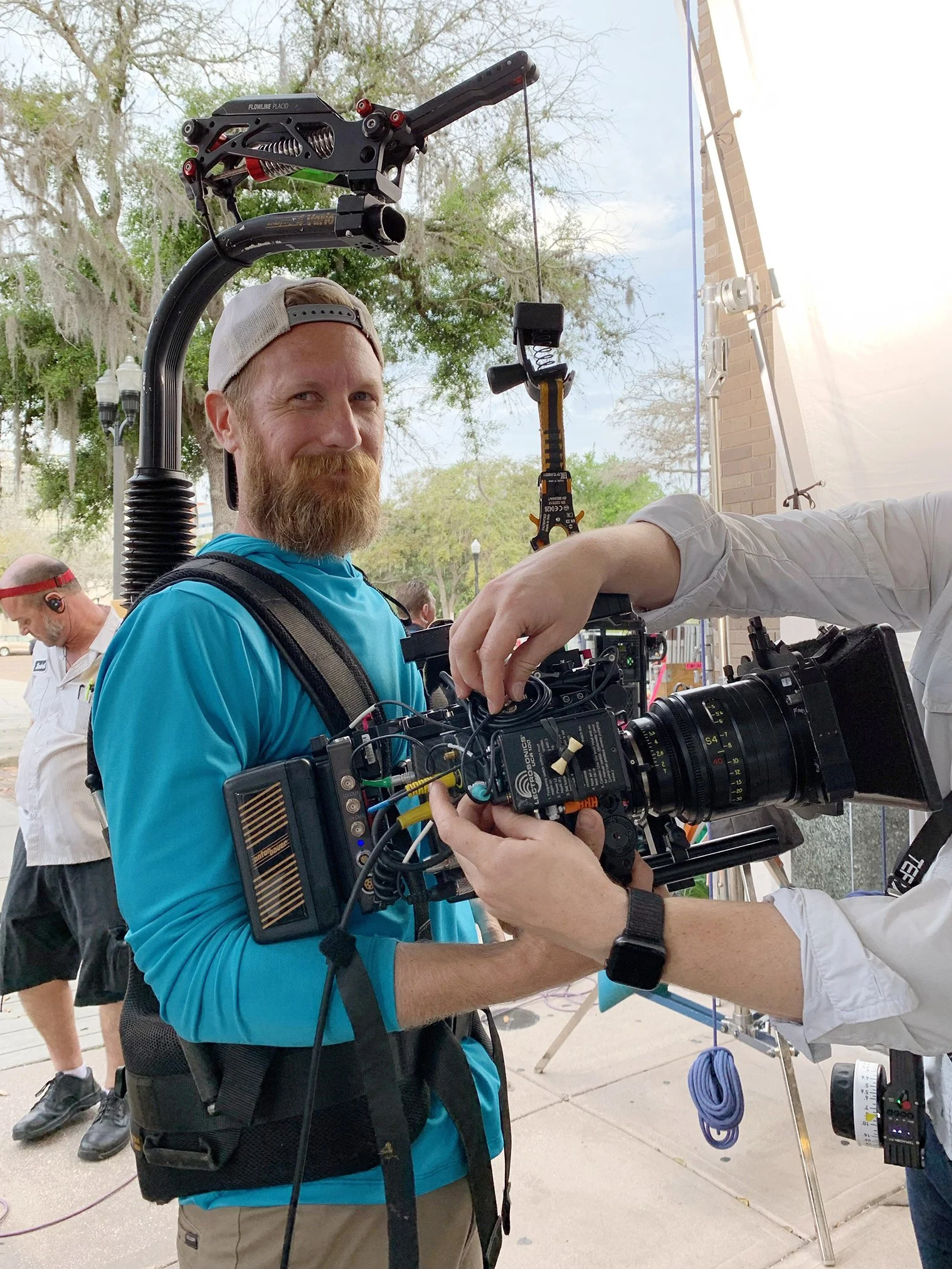 A man with a beard and cap being filmed or photographed using a professional camera and equipment outdoors, with another person adjusting the camera settings.