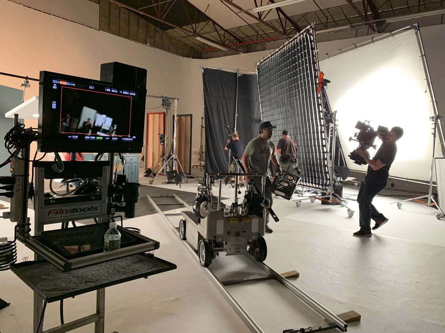 Film set with crew members preparing equipment, large lighting setup, camera, and monitor, indicating a film or television production in progress.