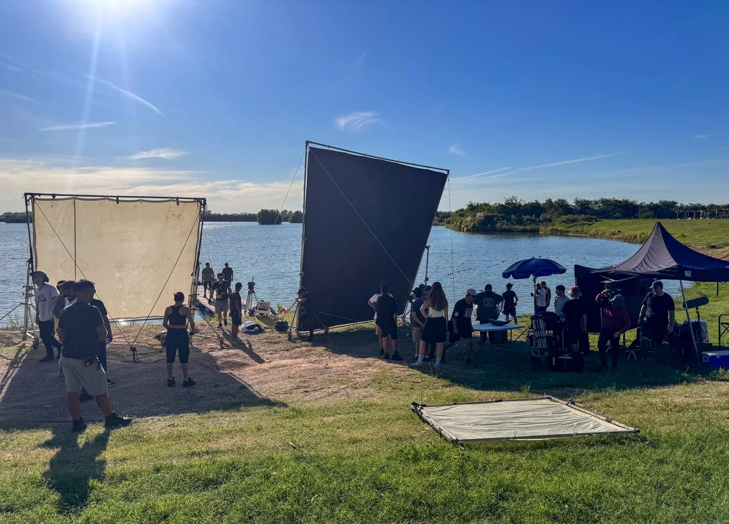People working on a film shoot near a body of water with large reflectors and a tent, under a sunny sky.