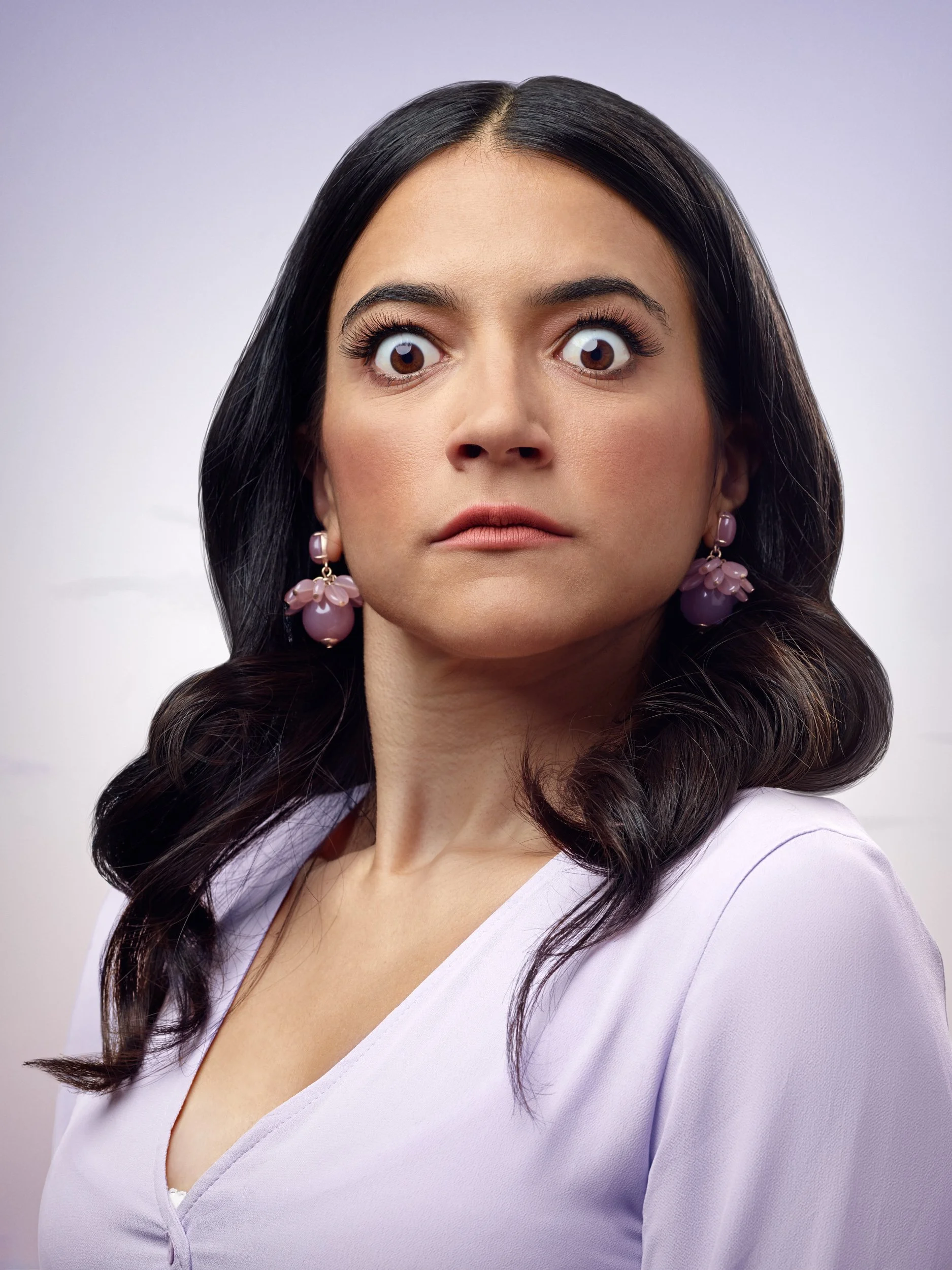 A woman with long dark hair, wide eyes, and a serious expression, wearing purple earrings and a light purple top.