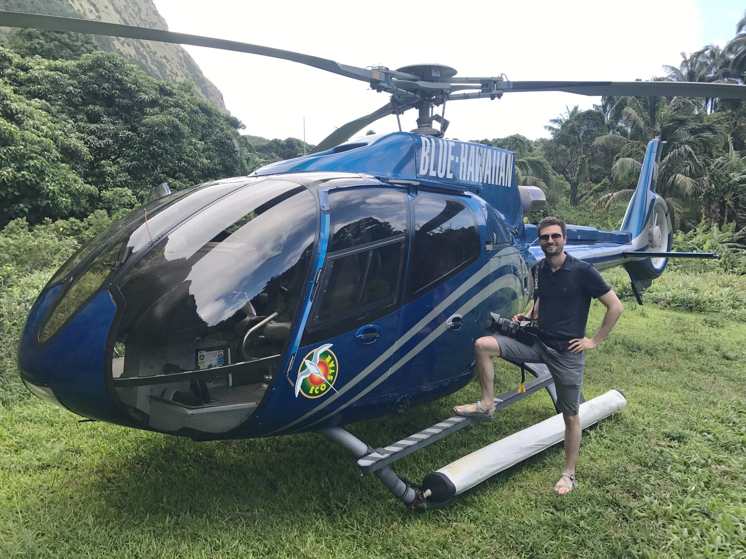 Man standing beside a blue helicopter labeled 'Blue Hawaii' on a grassy area surrounded by trees and mountains, holding a camera and wearing sunglasses, shorts, and sandals.
