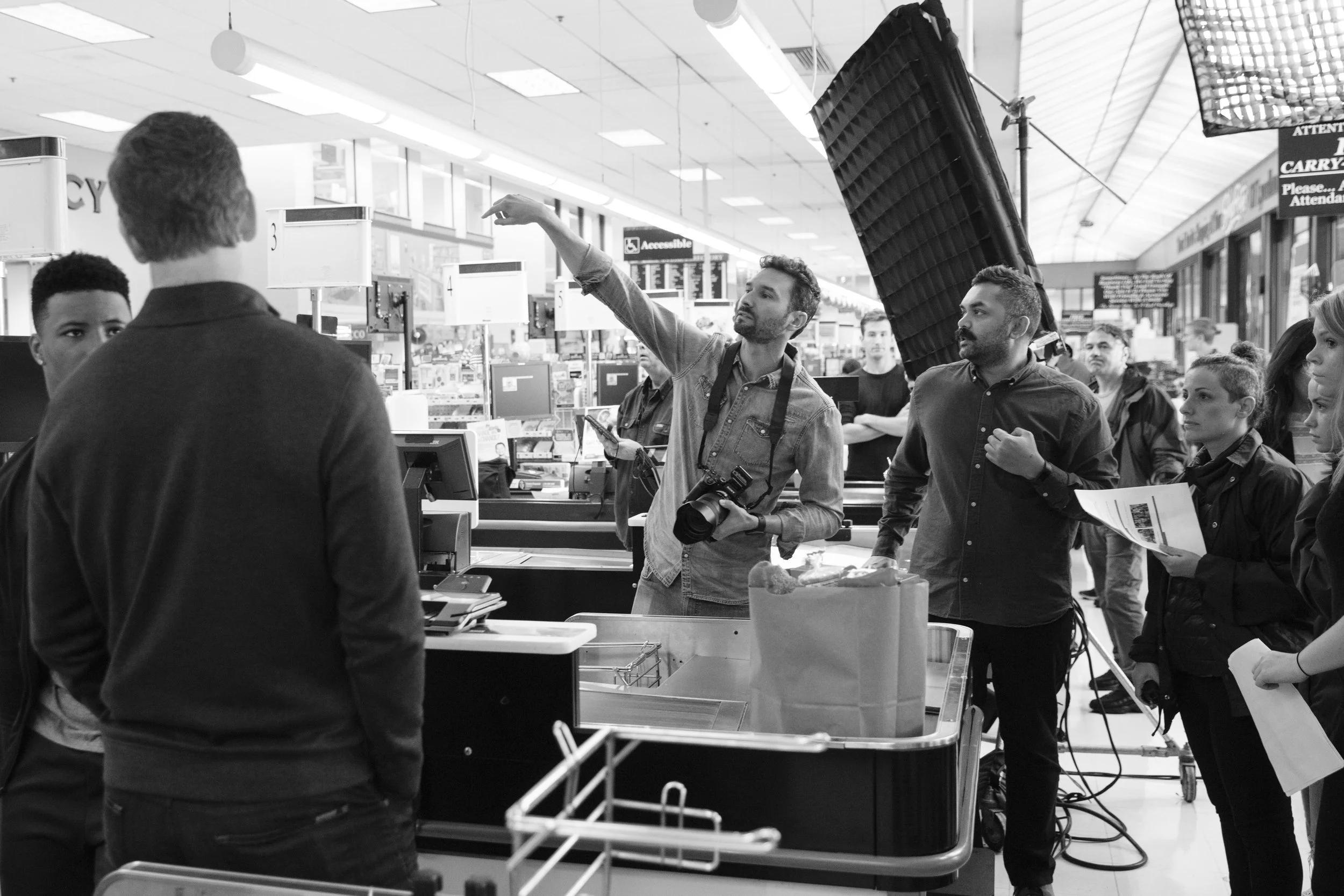 Filming crew setting up a scene at a supermarket checkout counter, with a camera operator adjusting a large light reflector and others preparing for a shoot.