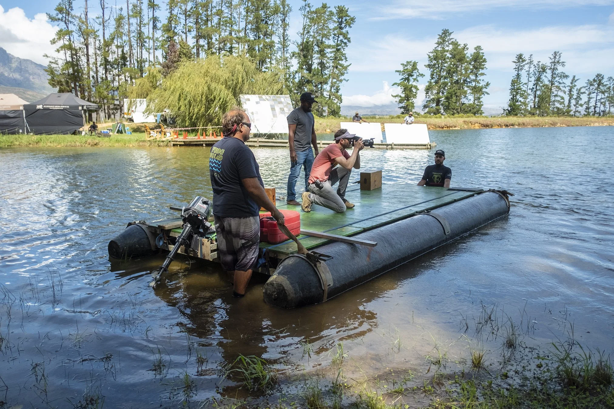 A group of people working on a flat platform on a river, with one person holding a camera, and others standing or sitting around, with trees, a dock, and tents in the background.