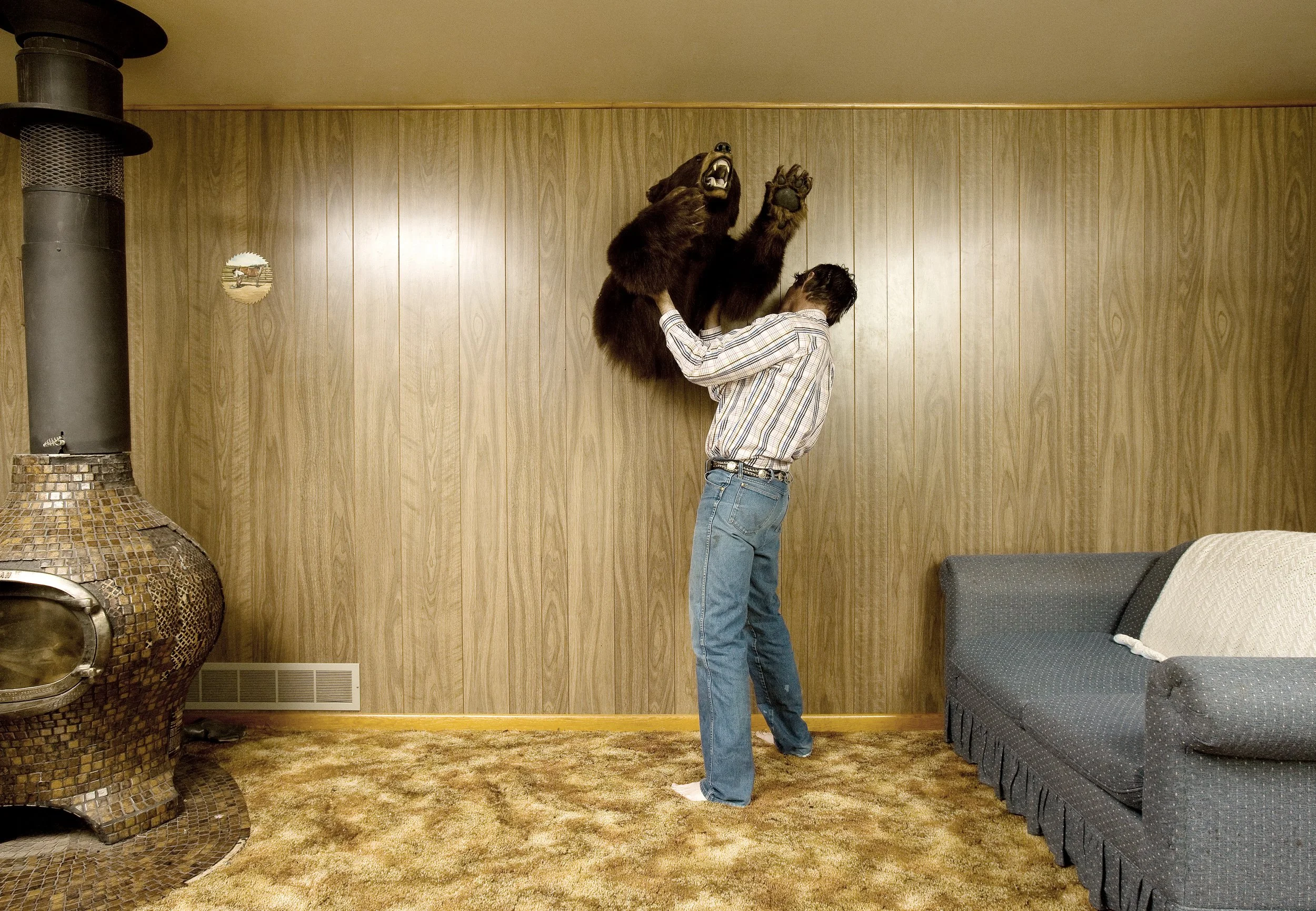 A person holding a mounted bear head on a wall in a room with wood-paneled walls and a patterned carpet.
