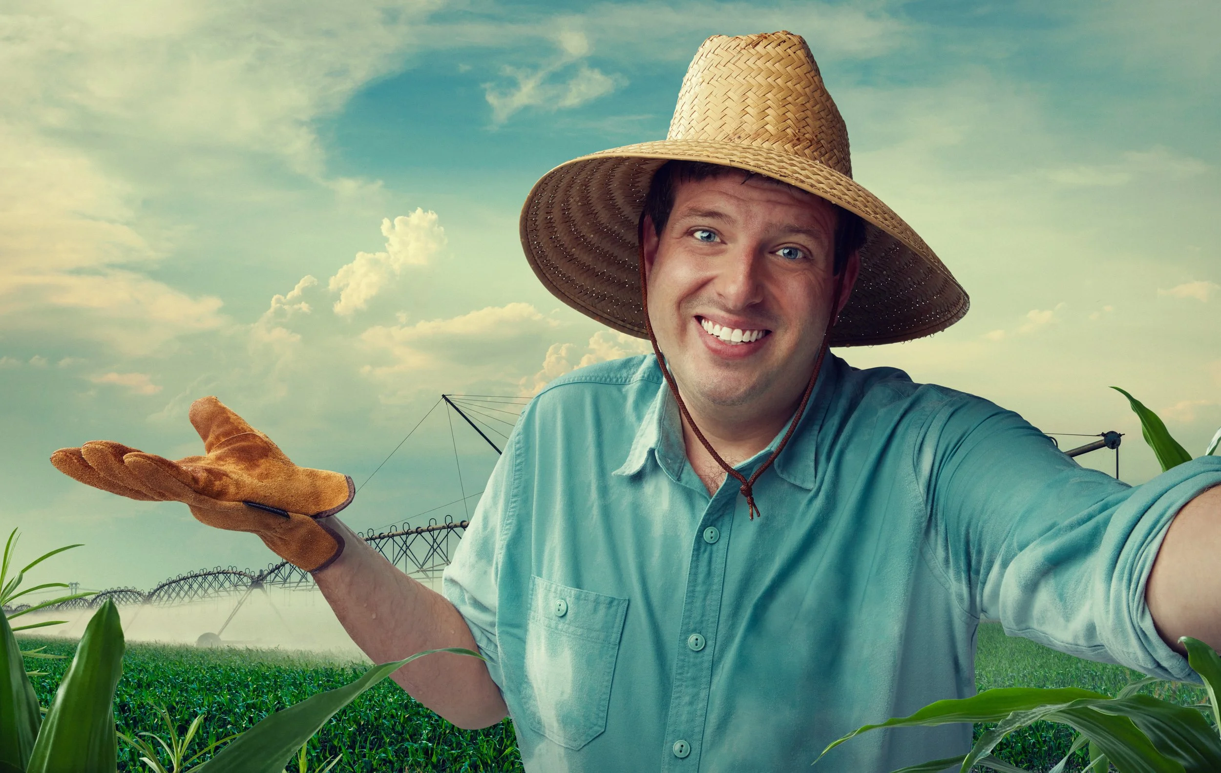 A man in a blue shirt and a large straw hat smiling and holding a gardening glove in a farm field with green plants and a sprinkling system in the background.