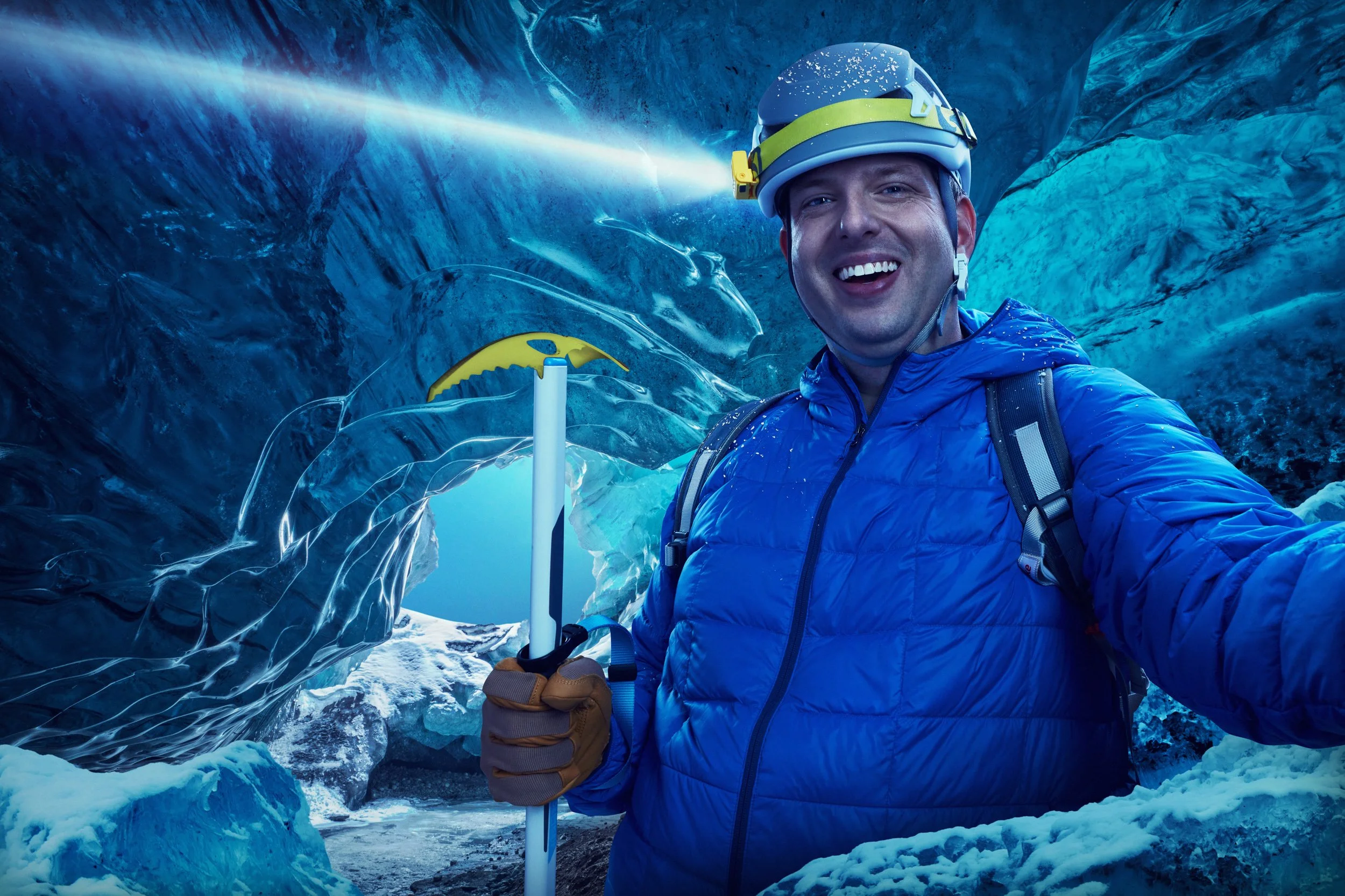 A man in a blue jacket and helmet taking a selfie inside an icy glacier cave, holding a white ice axe and smiling.