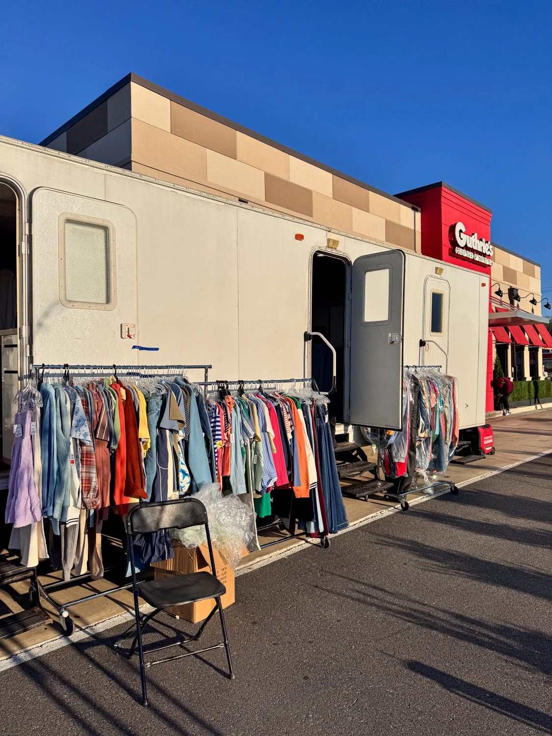 Outdoor clothing sale with racks of clothes set up outside a store, with a mobile trailer in the parking lot and a store building in the background.