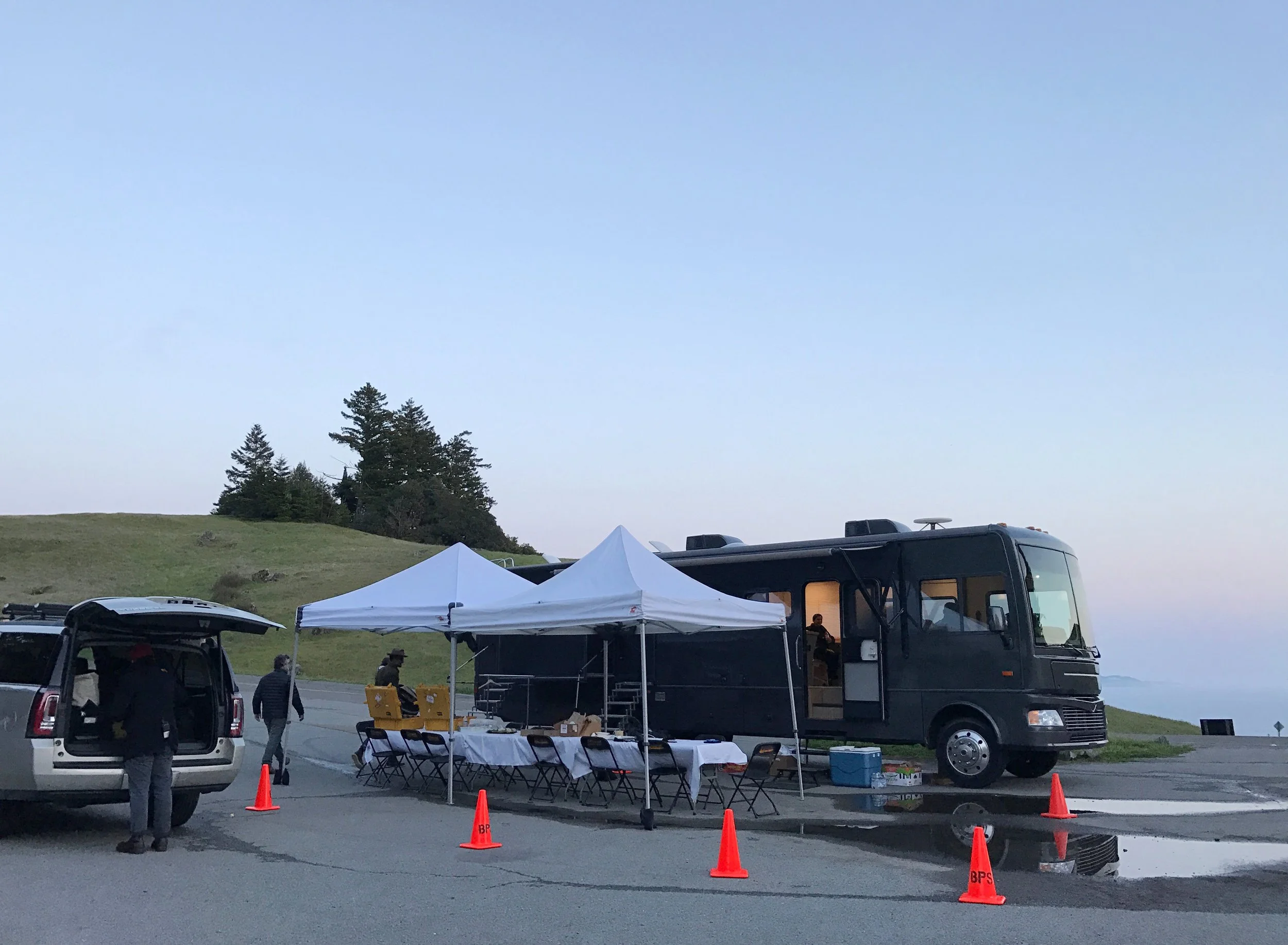 A mobile command unit or office setup with a black RV, white canopy tents, tables, chairs, and orange traffic cones in an outdoor parking area near a grassy hill and trees at dusk.