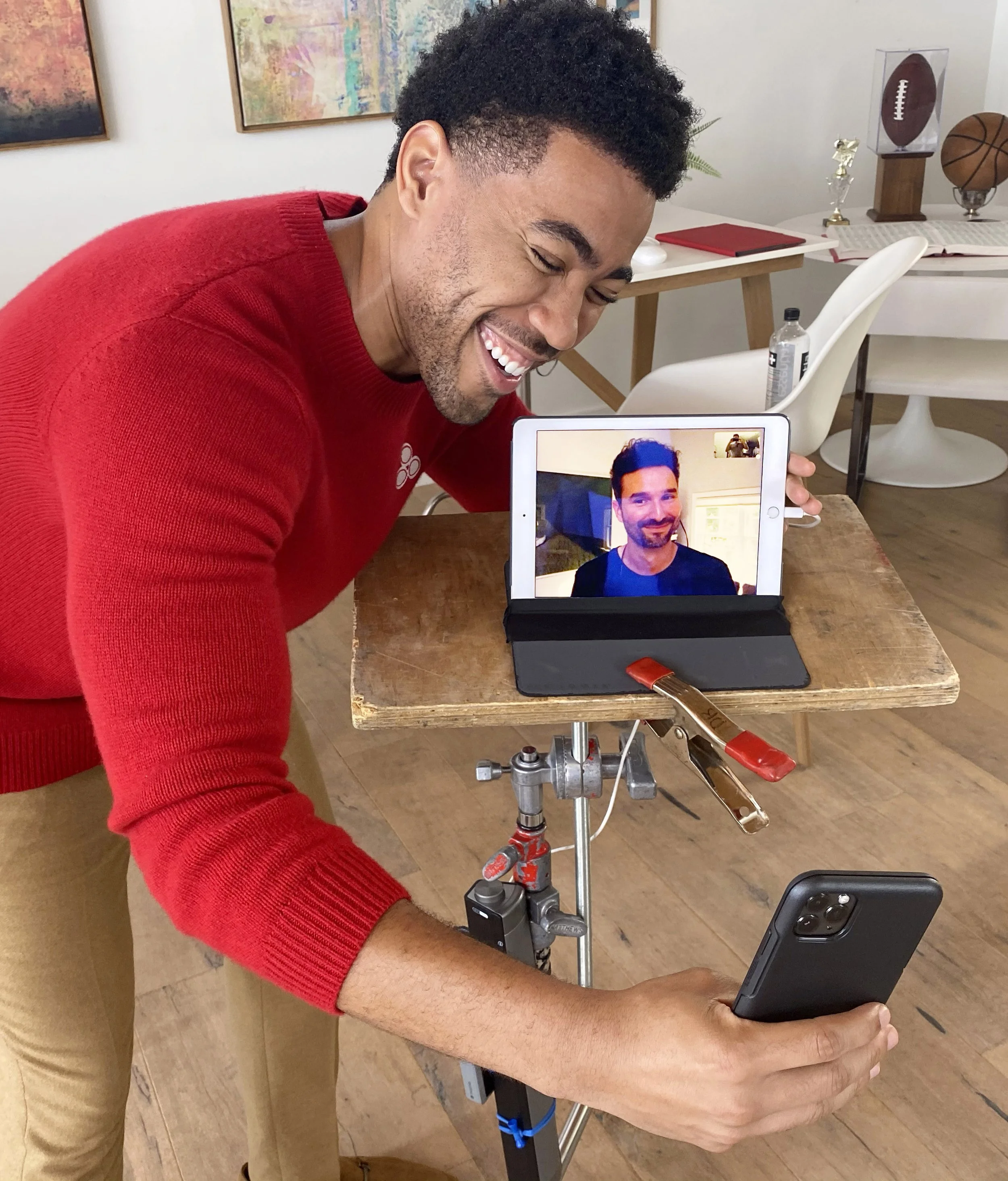 Man smiling and holding a smartphone in front of a video call on a tablet, which shows another man smiling. The setup includes a wooden table, a professional camera rig, and a bright, modern room with artwork, chairs, and sports memorabilia in the ba