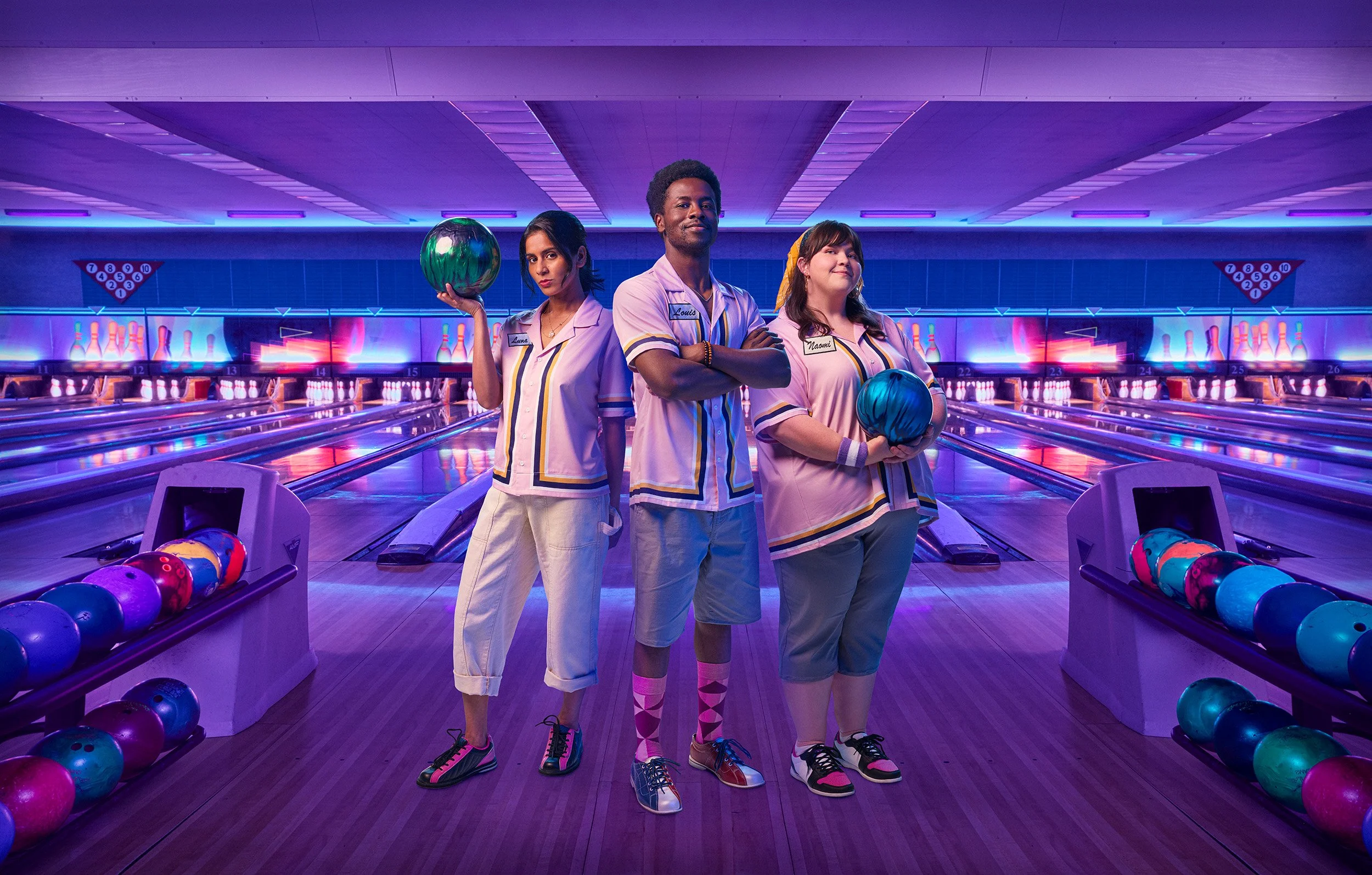Three young adults standing in a bowling alley holding bowling balls, wearing colorful retro-style bowling shirts, with lanes and neon lights in the background.