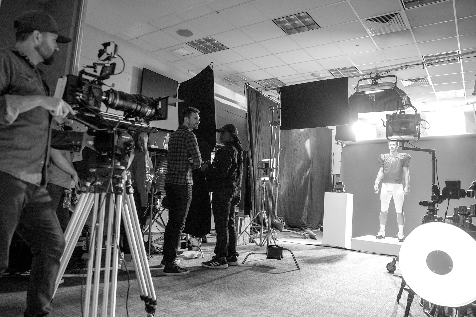 Film set with a camera operator, crew members, and a model standing on a white platform in front of a black background, with lighting equipment overhead.