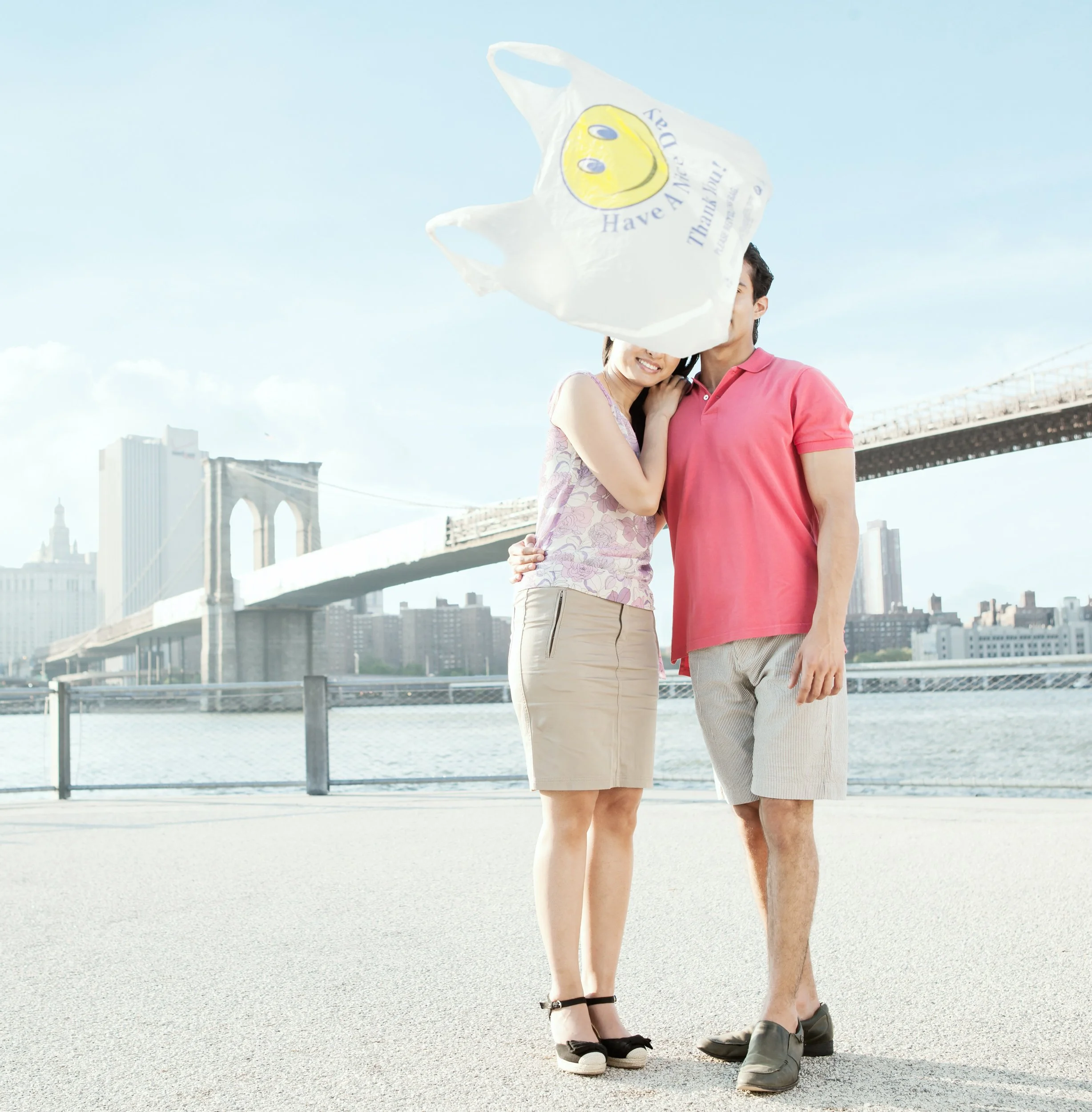 A couple standing close together by the waterfront under the Brooklyn Bridge in New York City, with the woman holding a white plastic bag with a yellow smiley face and the words 'Have A Nice Day!'