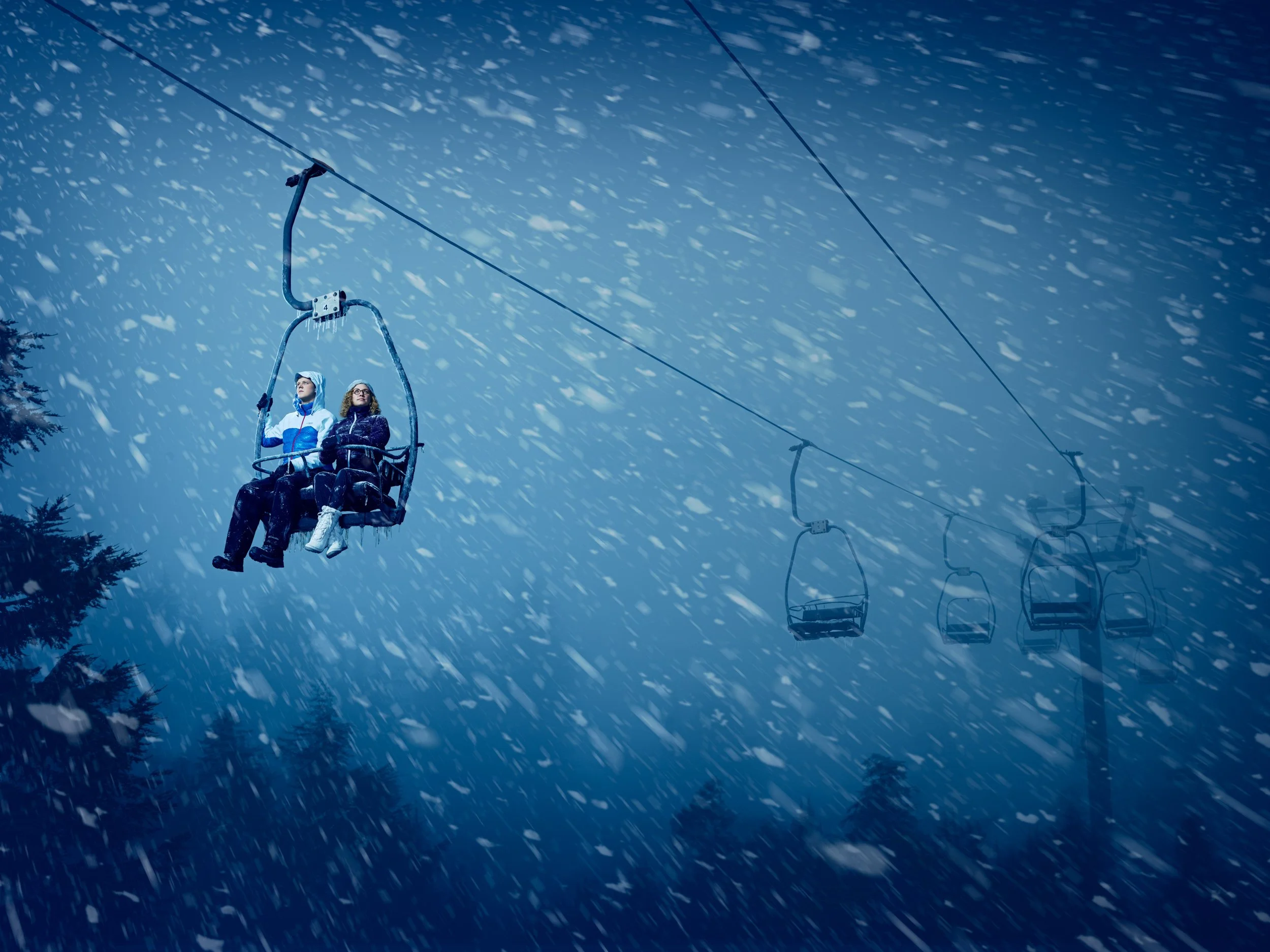 Two people riding a ski lift during heavy snowfall at night, with trees visible in the background.