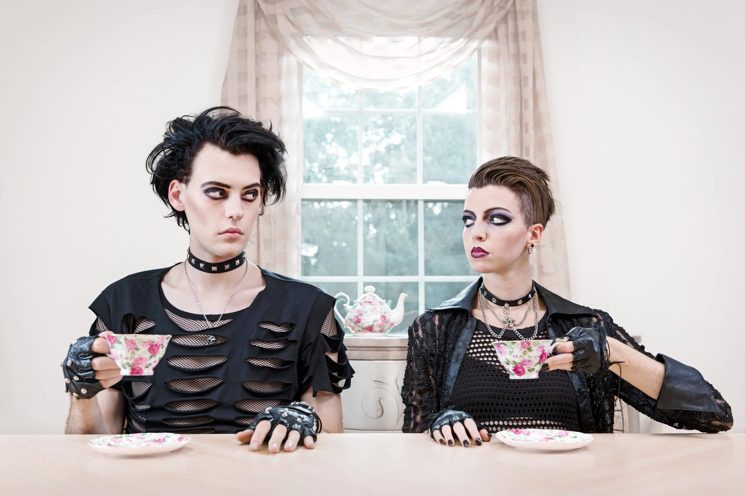 Two individuals with punk/goth style hair and makeup sitting at a table, holding floral teacups with matching saucers, in a room with a window and curtains in the background.