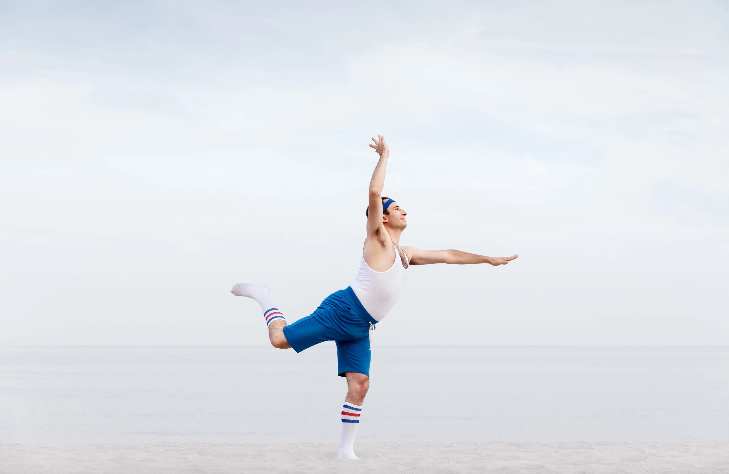 A man wearing blue shorts, a white tank top, striped knee-high socks, and a blue headband, is balancing on one leg on a beach while performing a yoga pose with one arm raised and the other extended forward. The background shows a cloudy sky and the o