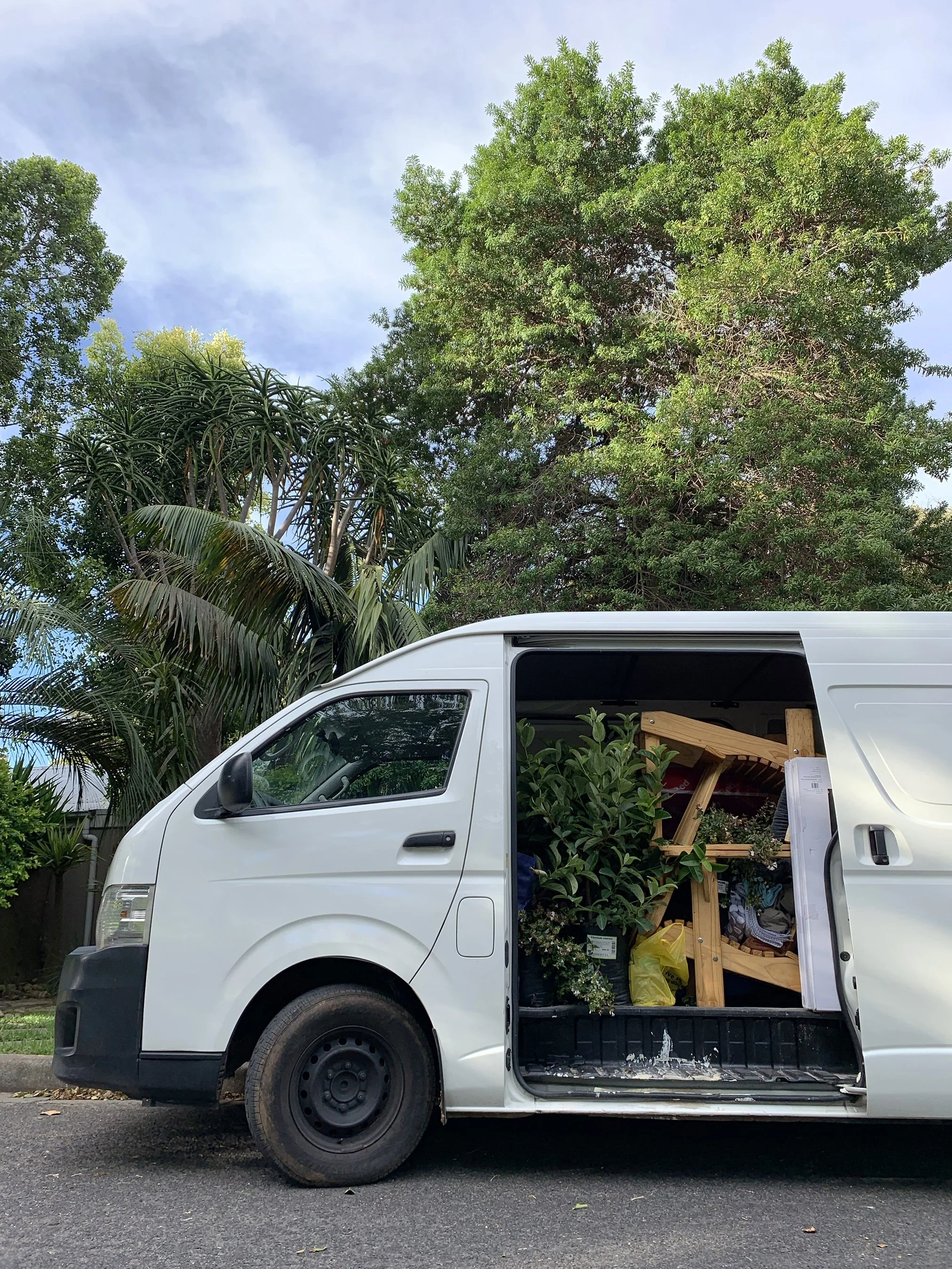White cargo van parked on street with open sliding door filled with plants, flowers, and wooden furniture, against a backdrop of large green trees and a partly cloudy sky.