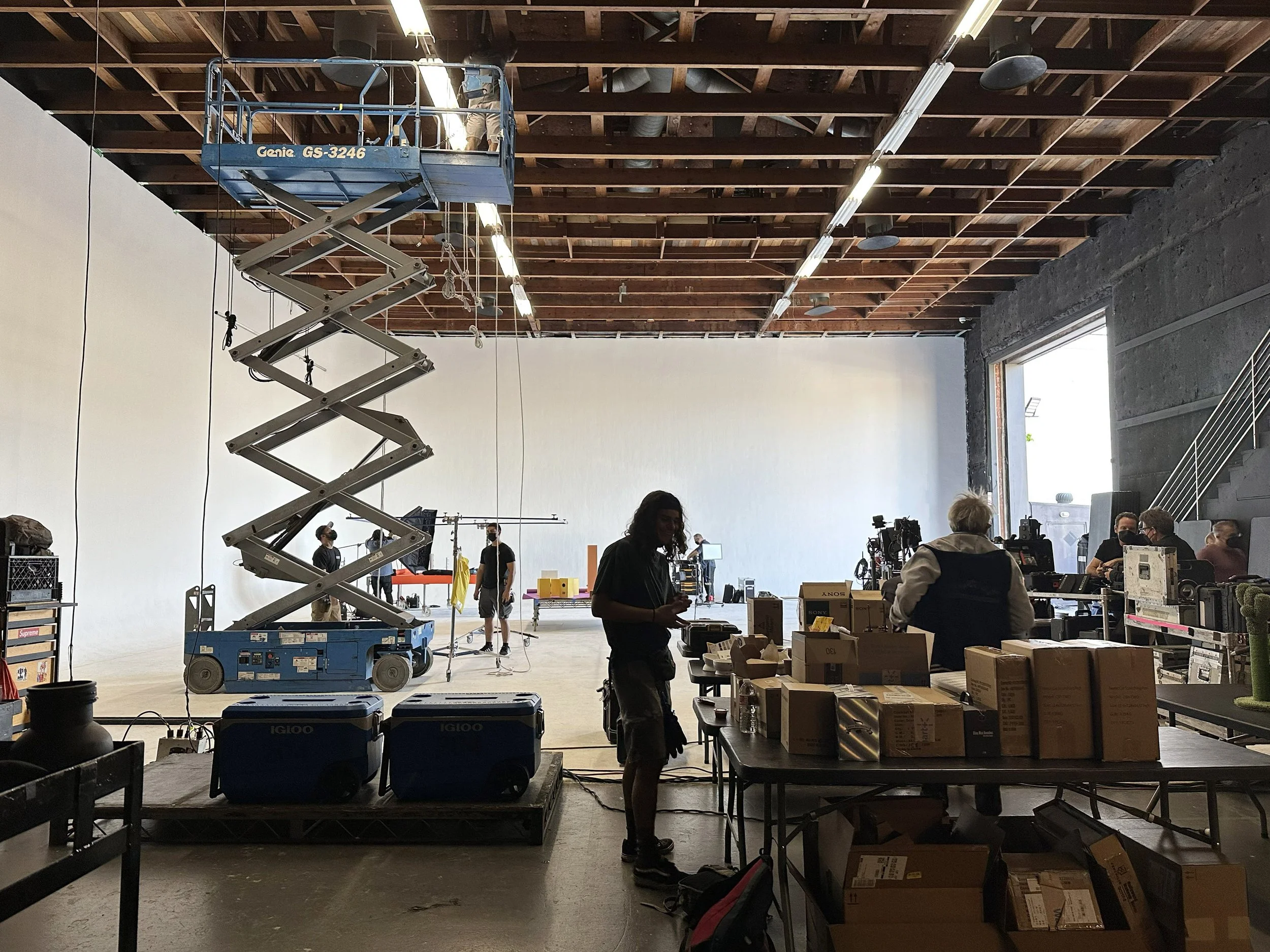 Film or photo shoot studio with a large open space, high ceiling, and crew members busy preparing. There is a scissor lift, camera equipment, and supplies, with a bright open doorway letting in natural light.