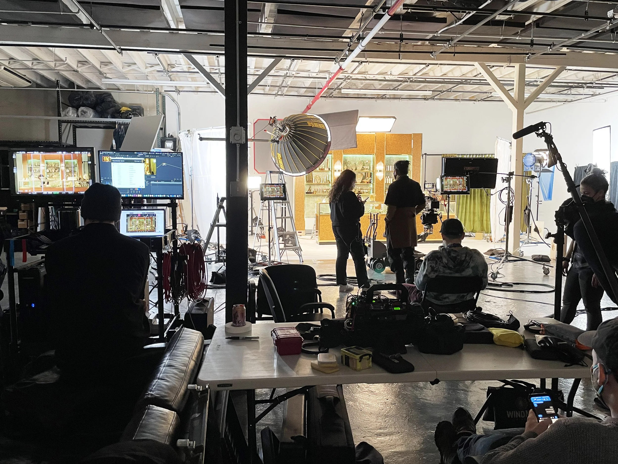 Behind-the-scenes view of a filming set with crew members, cameras, and lighting equipment, and a colorful display cabinet in the background.