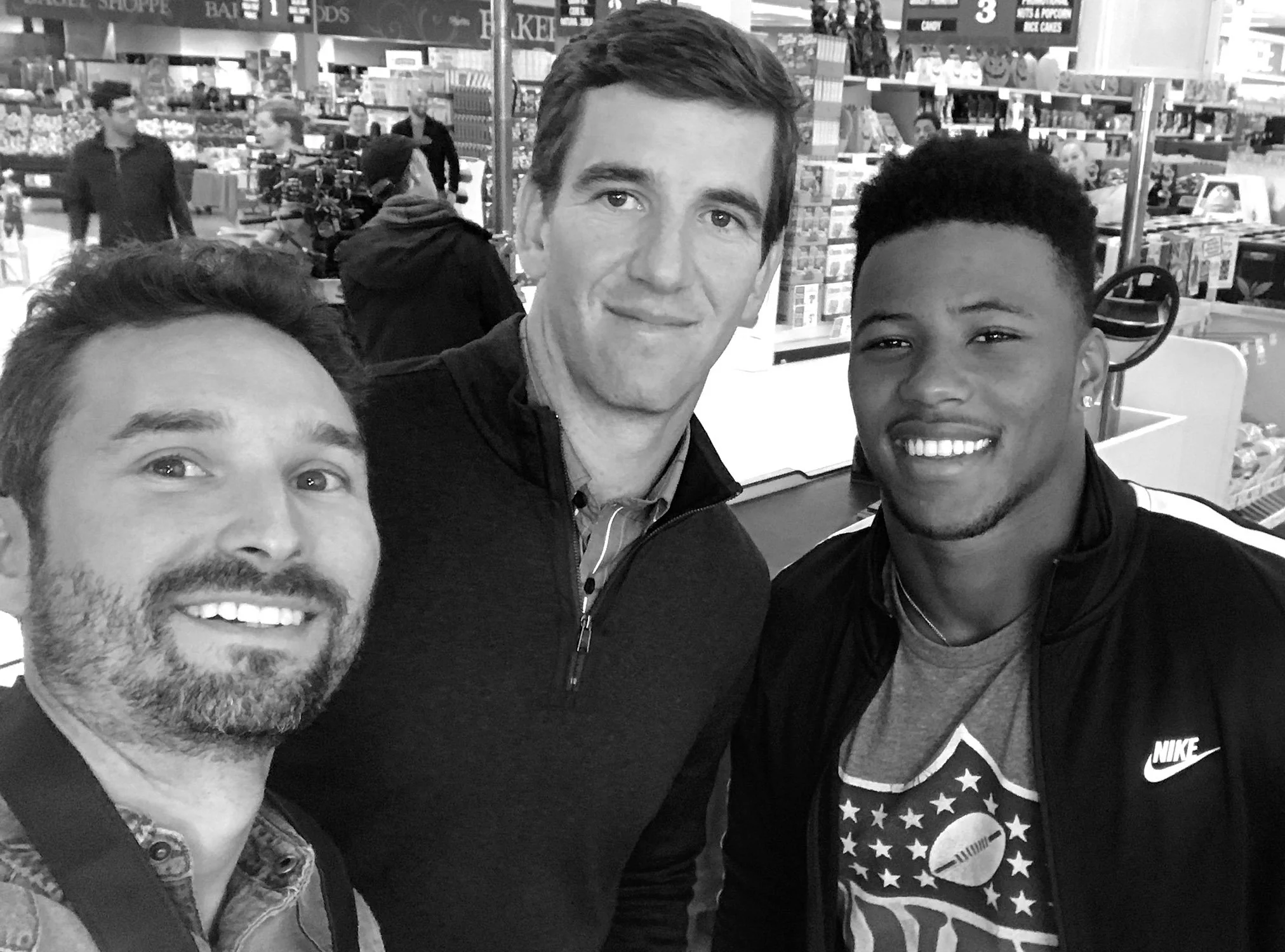 Three men taking a selfie inside a store or shopping mall, with shelves of products and other shoppers in the background.