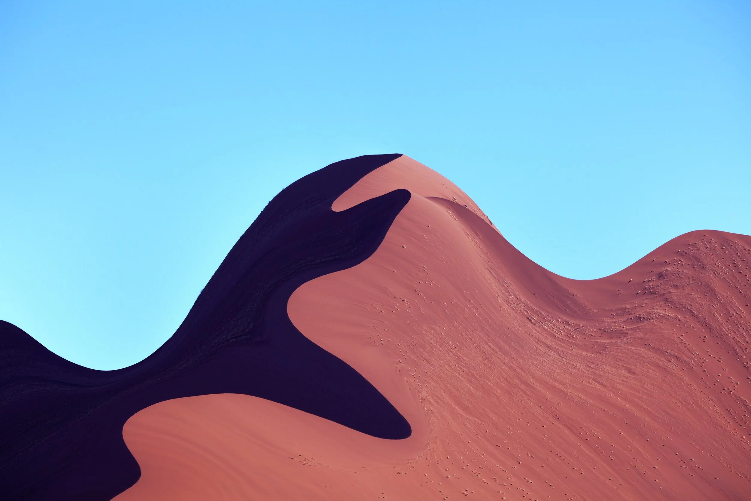 A large desert sand dune with a dark shadow on one side, set against a clear blue sky.