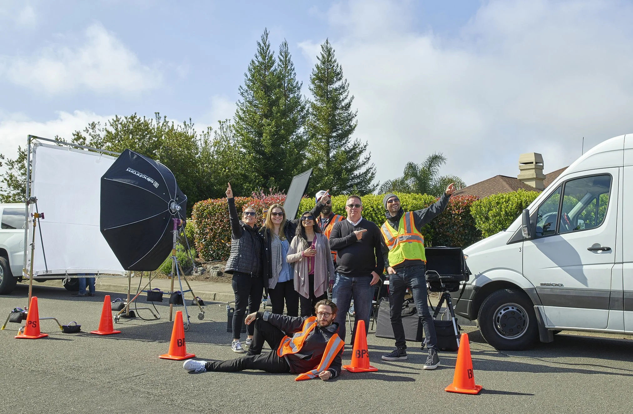 Group of people on a film set, with a white van, safety cones, lighting equipment, and trees in the background, all appearing to be having fun and posing for a photo.