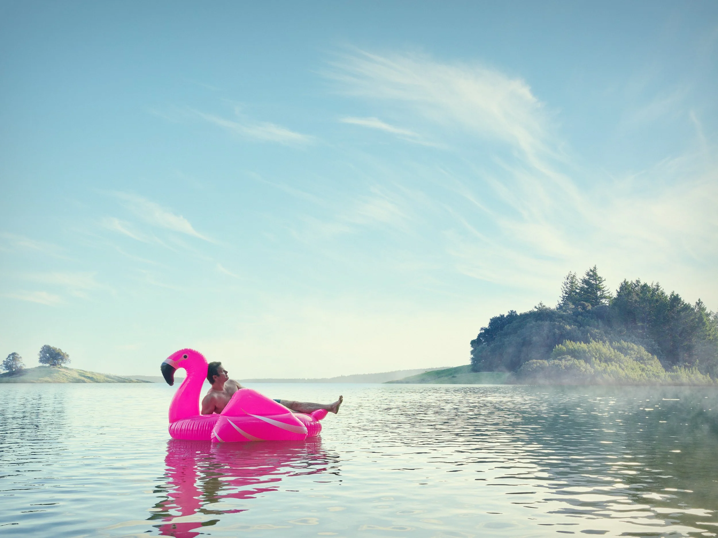 A man relaxing on an inflatable pink flamingo in a calm lake, with a clear sky and distant trees and hills in the background.