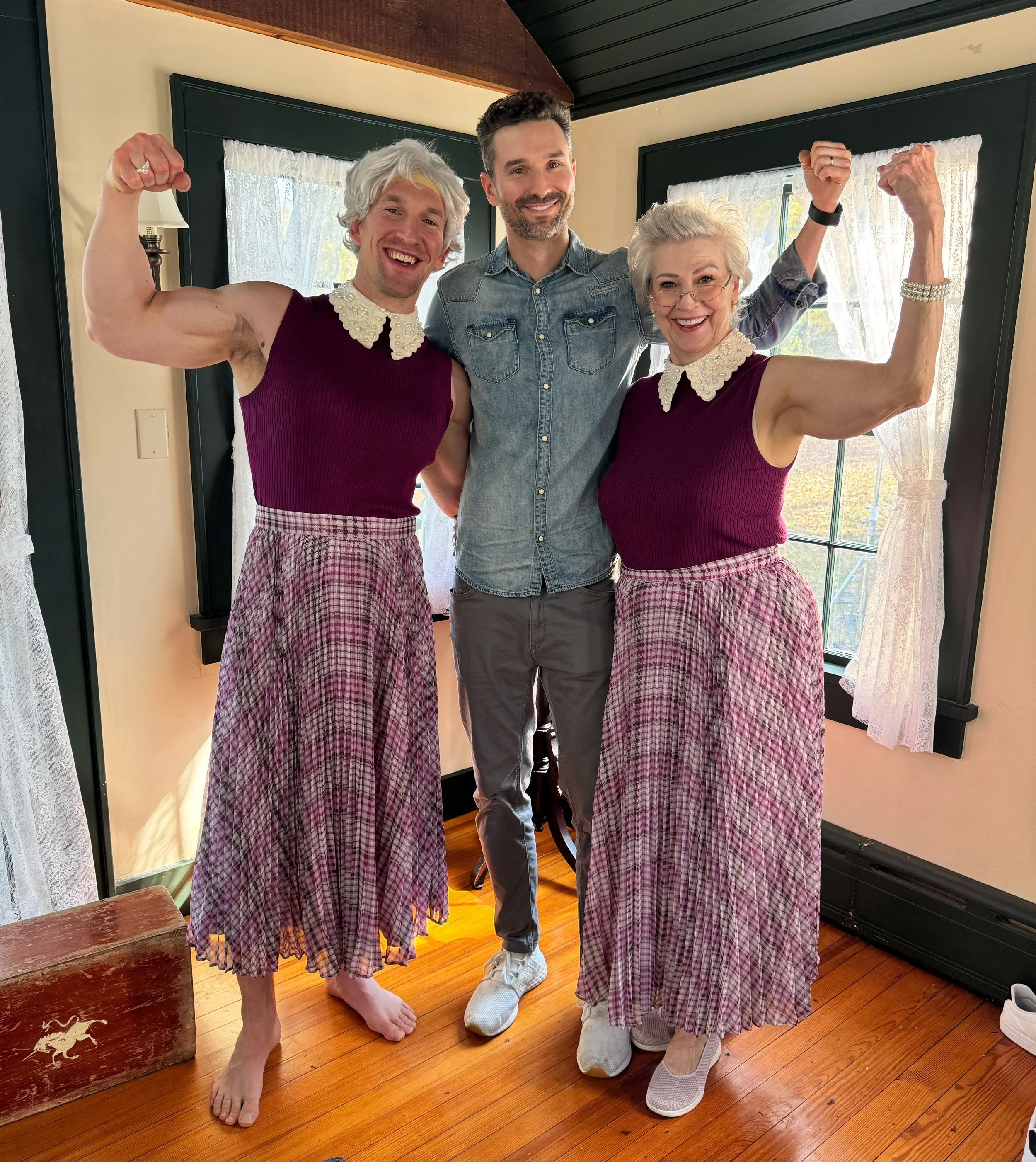 Two elderly women with short white hair, wearing matching sleeveless purple tops with white lace collars and pink checkered long skirts, are flexing their muscles and smiling. A middle-aged man with short brown hair, a beard, wearing a denim shirt an