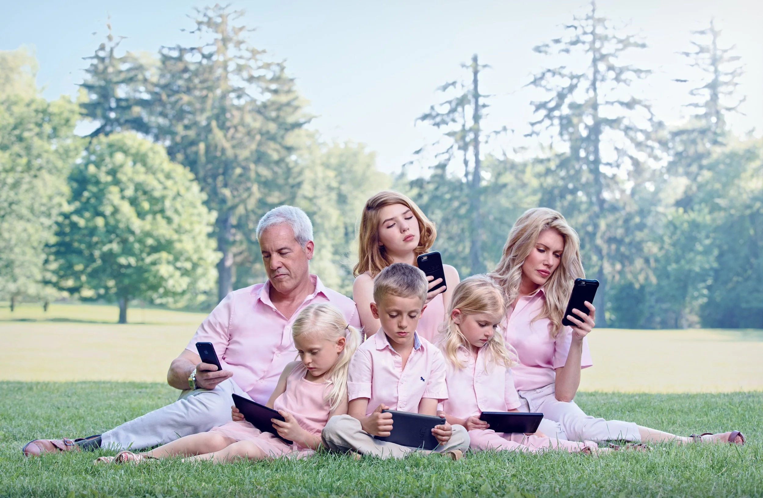 Family of six sitting on grass in park, all looking at smartphones or tablets