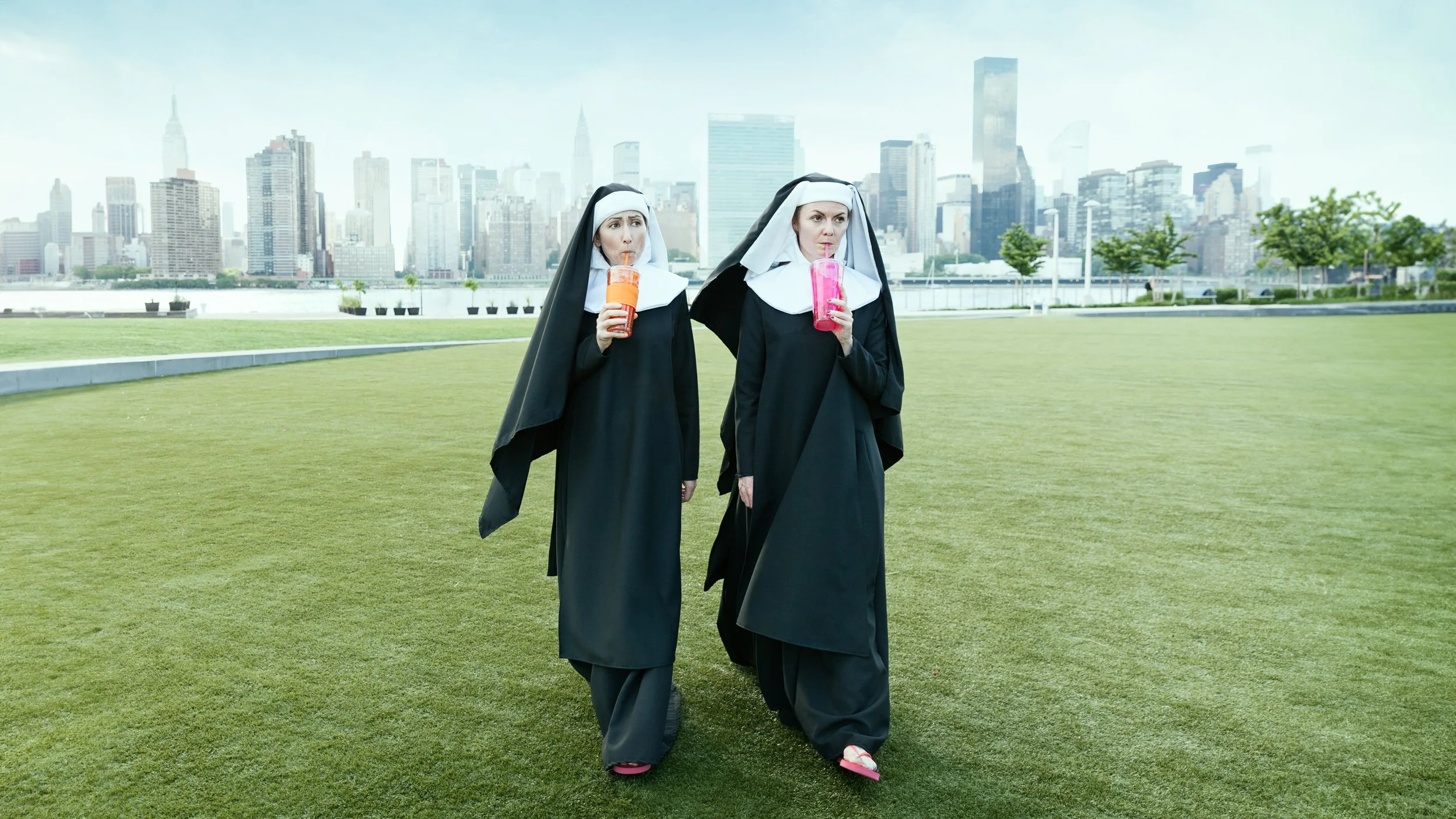 Two women dressed as nuns walking on a grassy field and drinking sodas, with a city skyline featuring tall buildings in the background.