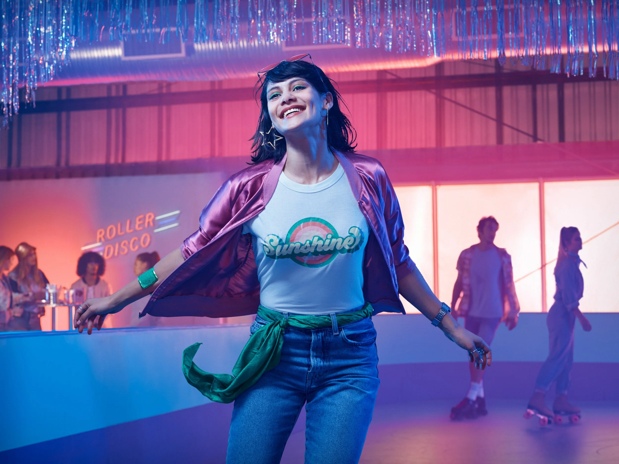 A woman roller skating indoors with a big smile, wearing a T-shirt that says 'Sunshine,' a shiny pink jacket, blue jeans, and large star-shaped earrings. There are other people roller skating in the background and a neon sign that says 'Roller Disco.