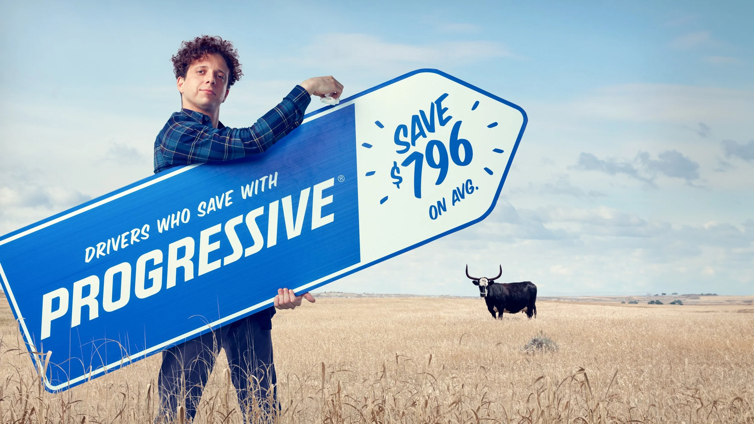 A man holding a large blue and white sign that reads 'Drivers who save with Progressive' and offers a $796 savings on average. The man is standing in a grassy field with a black buffalo in the background and a partly cloudy sky.