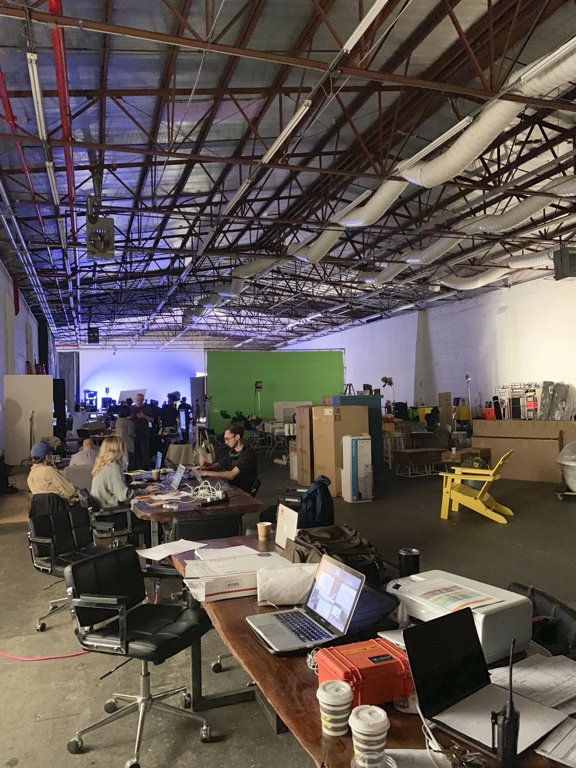 A large, indoor workspace or studio with high ceilings, exposed pipes and beams. There are multiple desks with laptops, papers, and coffee cups. Some people are working at the desks, while others are standing or moving around. In the background, ther