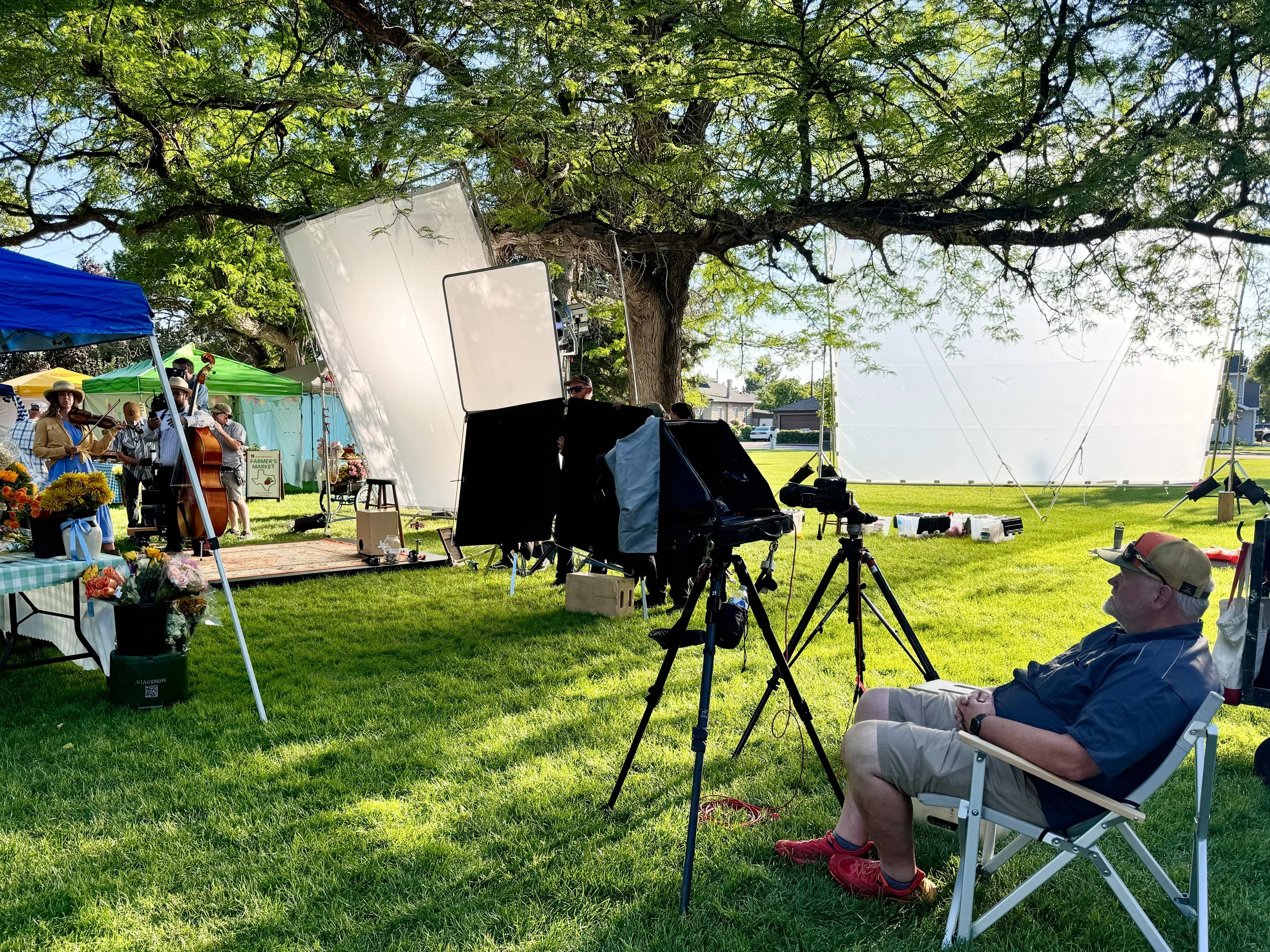Filming setup for outdoor event under a large tree, with a camera, tripods, black and white screens, and a man sitting in a chair, while a band plays music in the background.