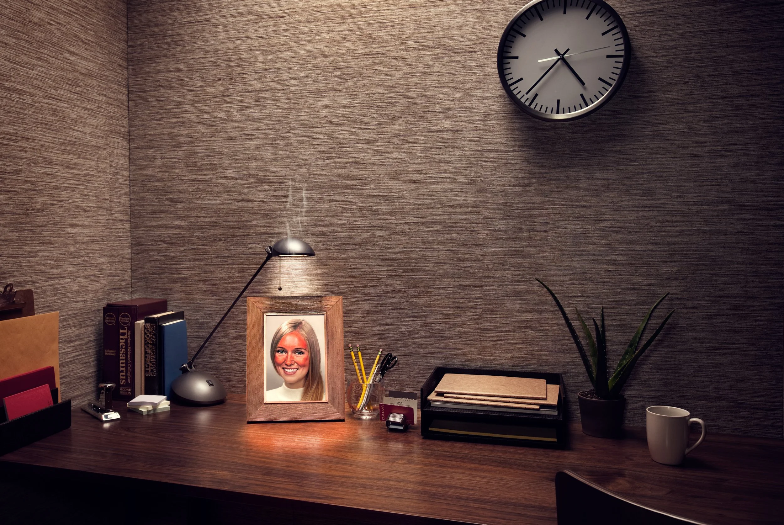 An office desk with a framed photo of a smiling woman, a desk lamp, books, office supplies, a potted plant, a coffee mug, and a wall clock showing 2:43.