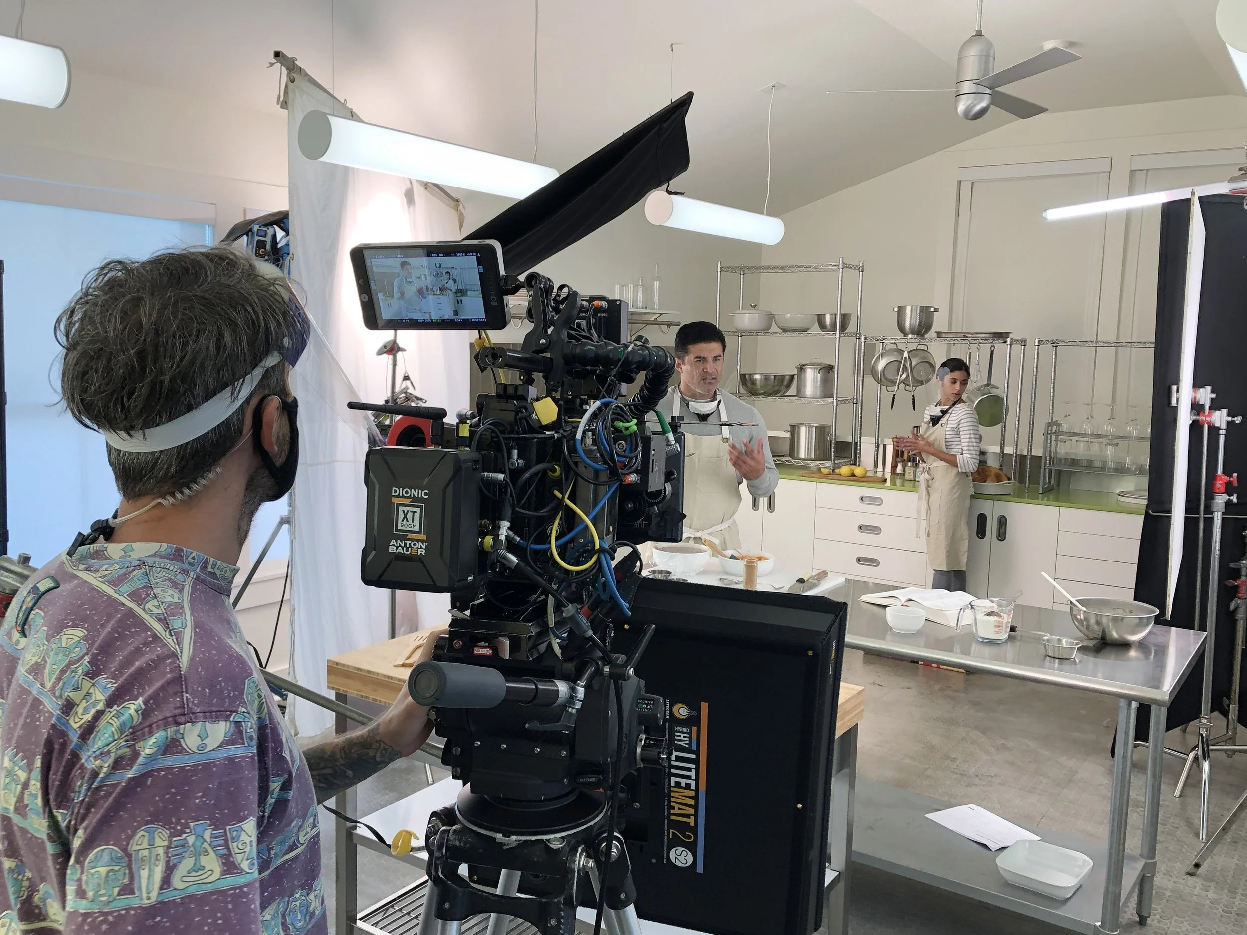 Filming a cooking show in a professional kitchen with two hosts, one man and one woman, while a crew member operates a camera.