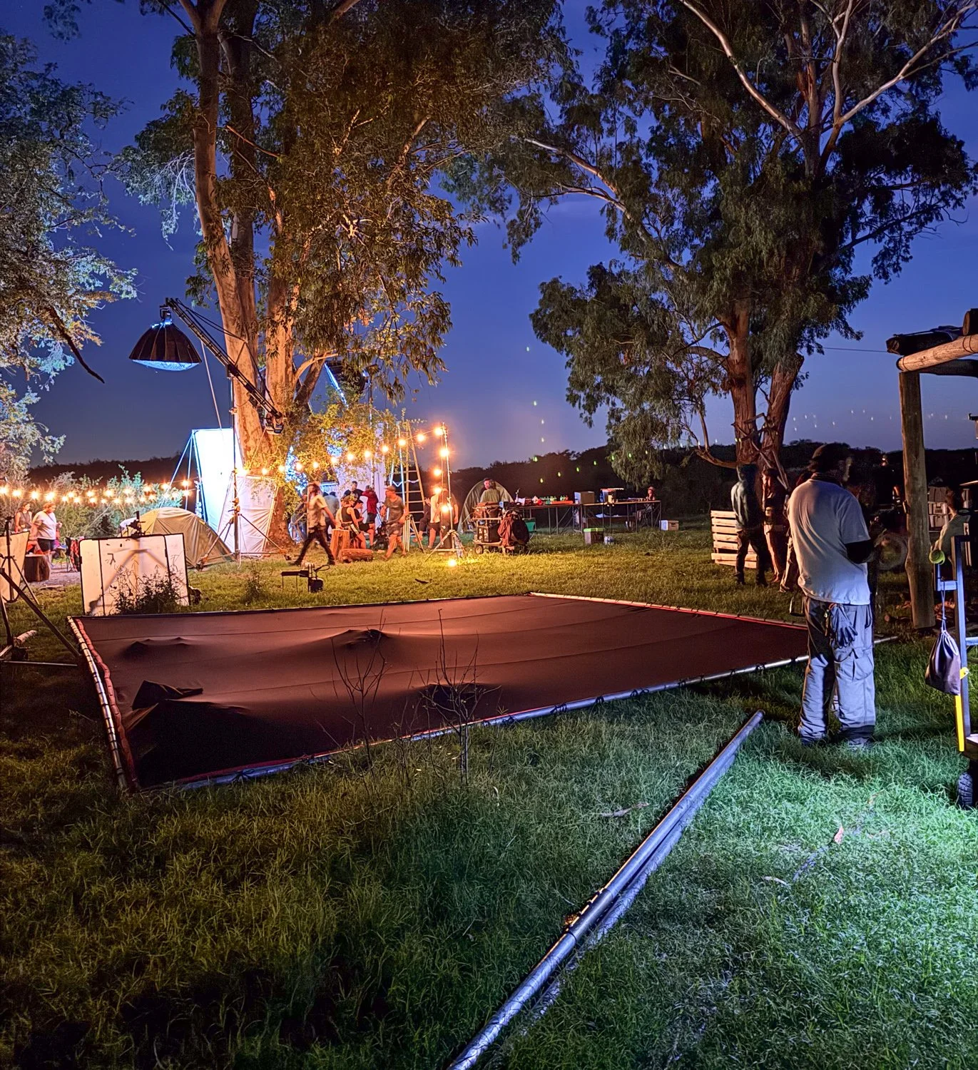 Nighttime outdoor gathering with string lights, people, tents, and equipment, possibly for an event or festival, near trees and open field.