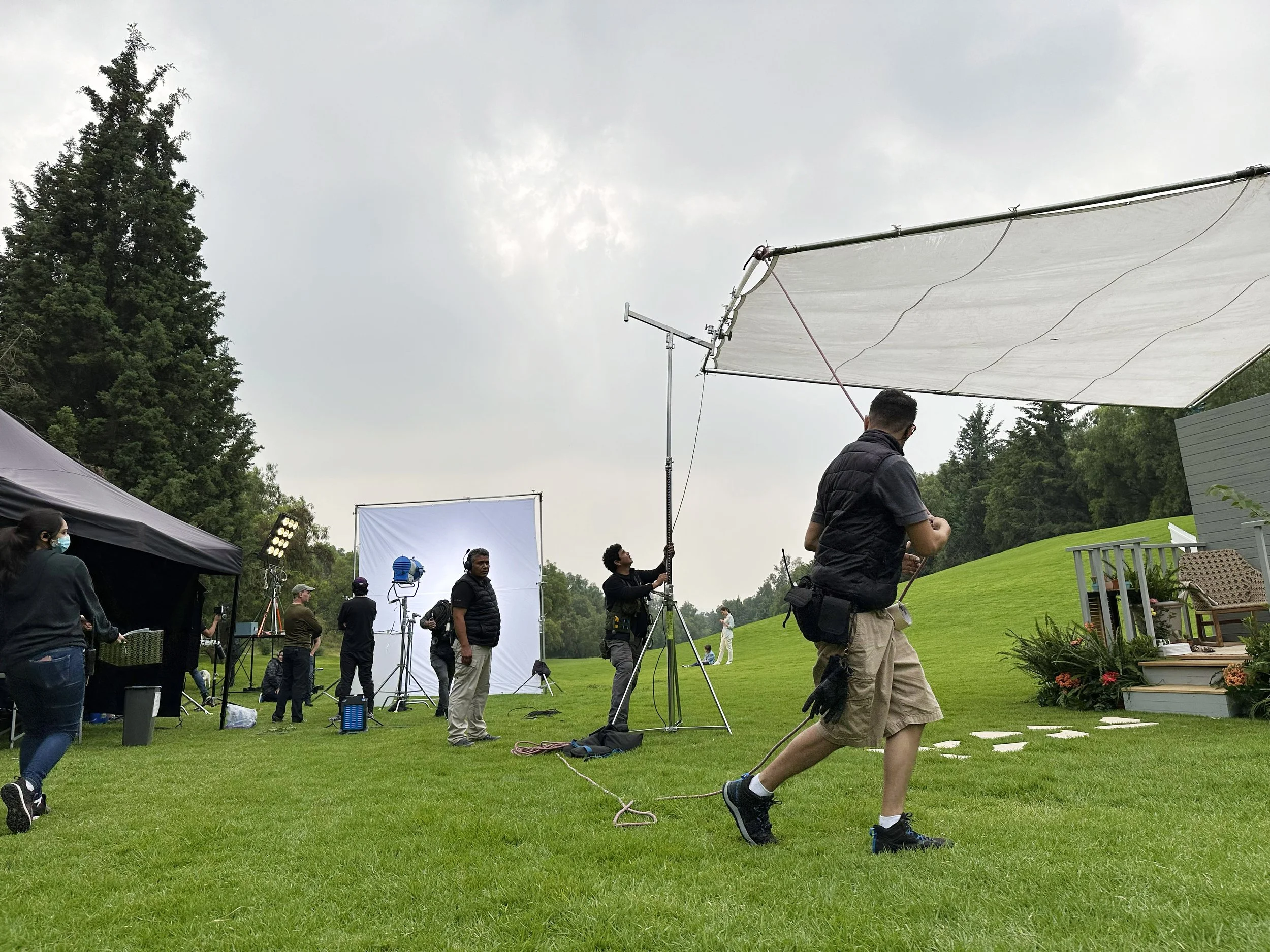 A film crew setting up equipment outdoors on a lawn with a hill and trees in the background, preparing for a shoot.