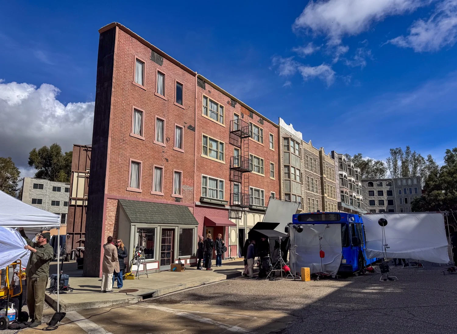 Behind-the-scenes view of a film or photo shoot with crew members and equipment on a city street, with a row of colorful apartment buildings in the background.