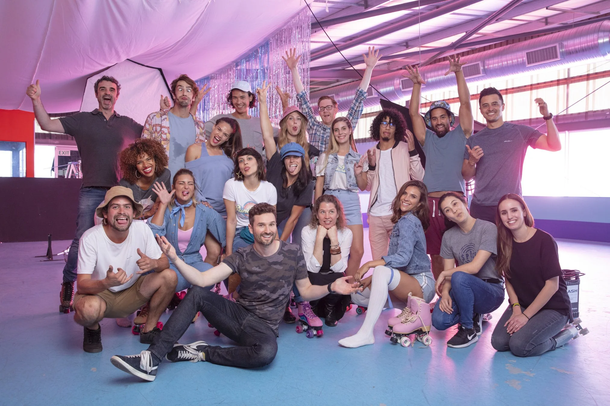 Group of young people at roller rink, smiling, some wearing roller skates, in bright colorful space with pink lighting.