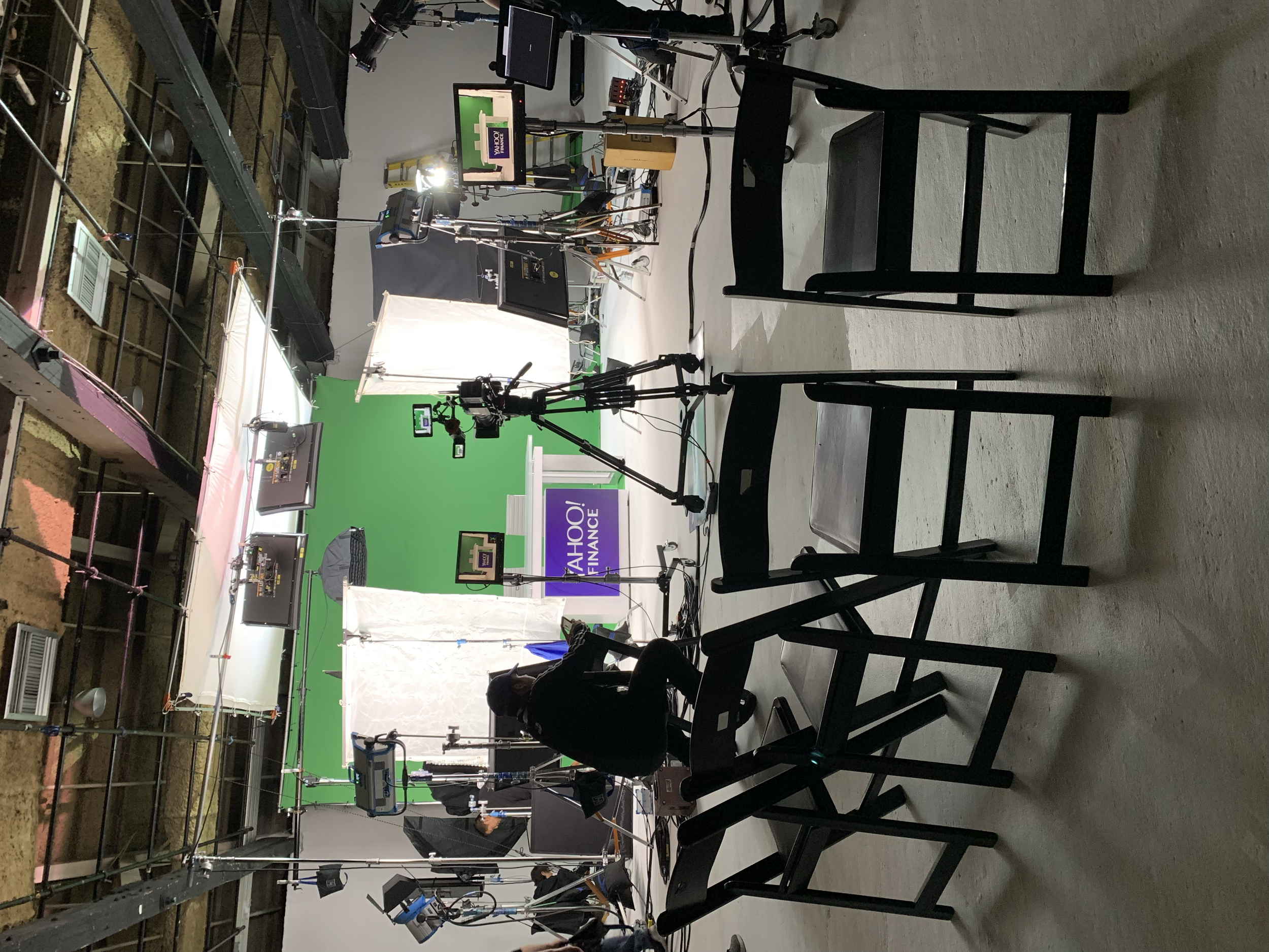 An indoor TV news studio with a green screen, cameras, monitors, lights, and crew members preparing for a broadcast.