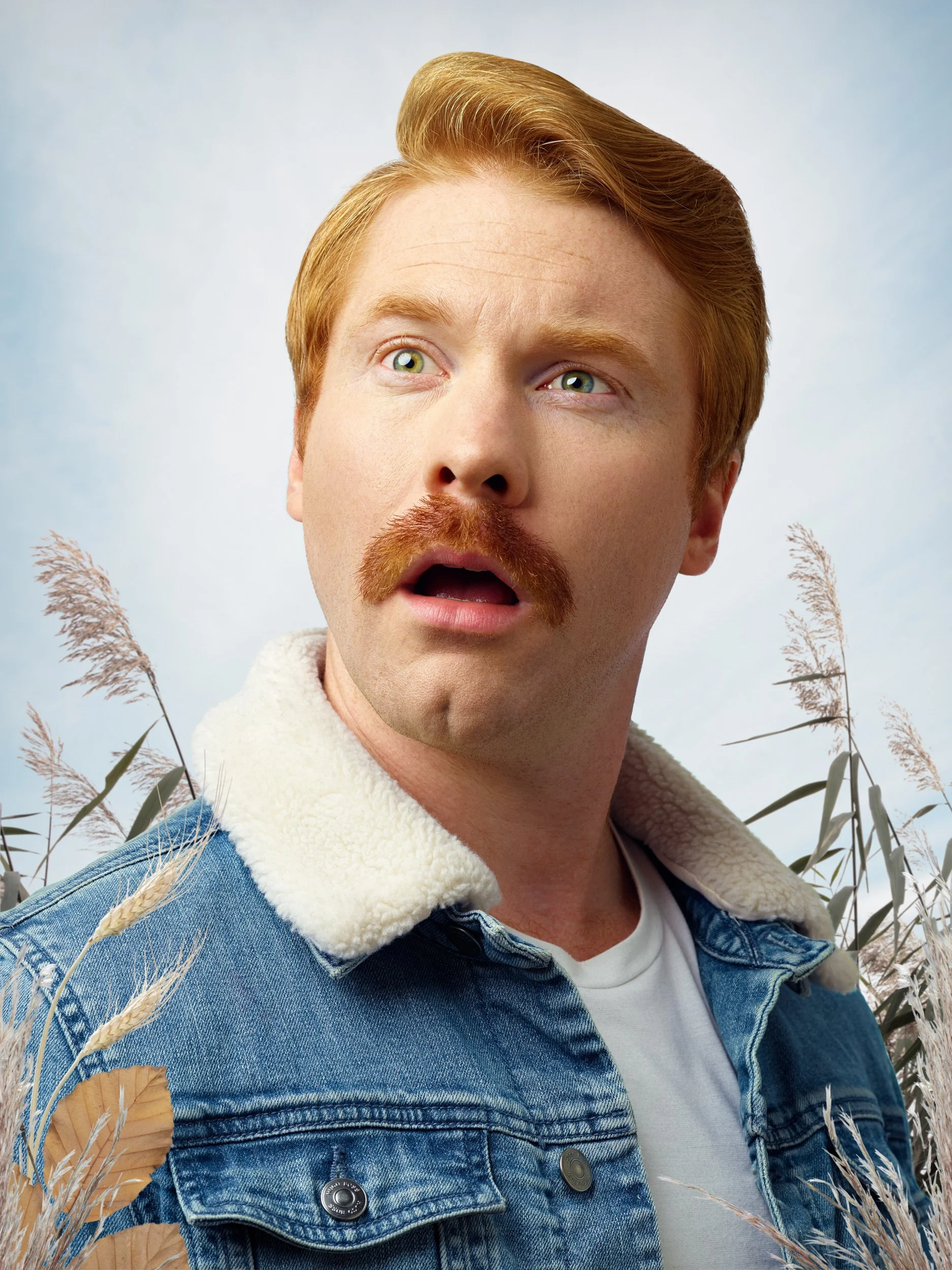 A man with red hair and mustache wearing a denim jacket with a fleece collar, standing outdoors in a field of tall grass, looking surprised or confused.