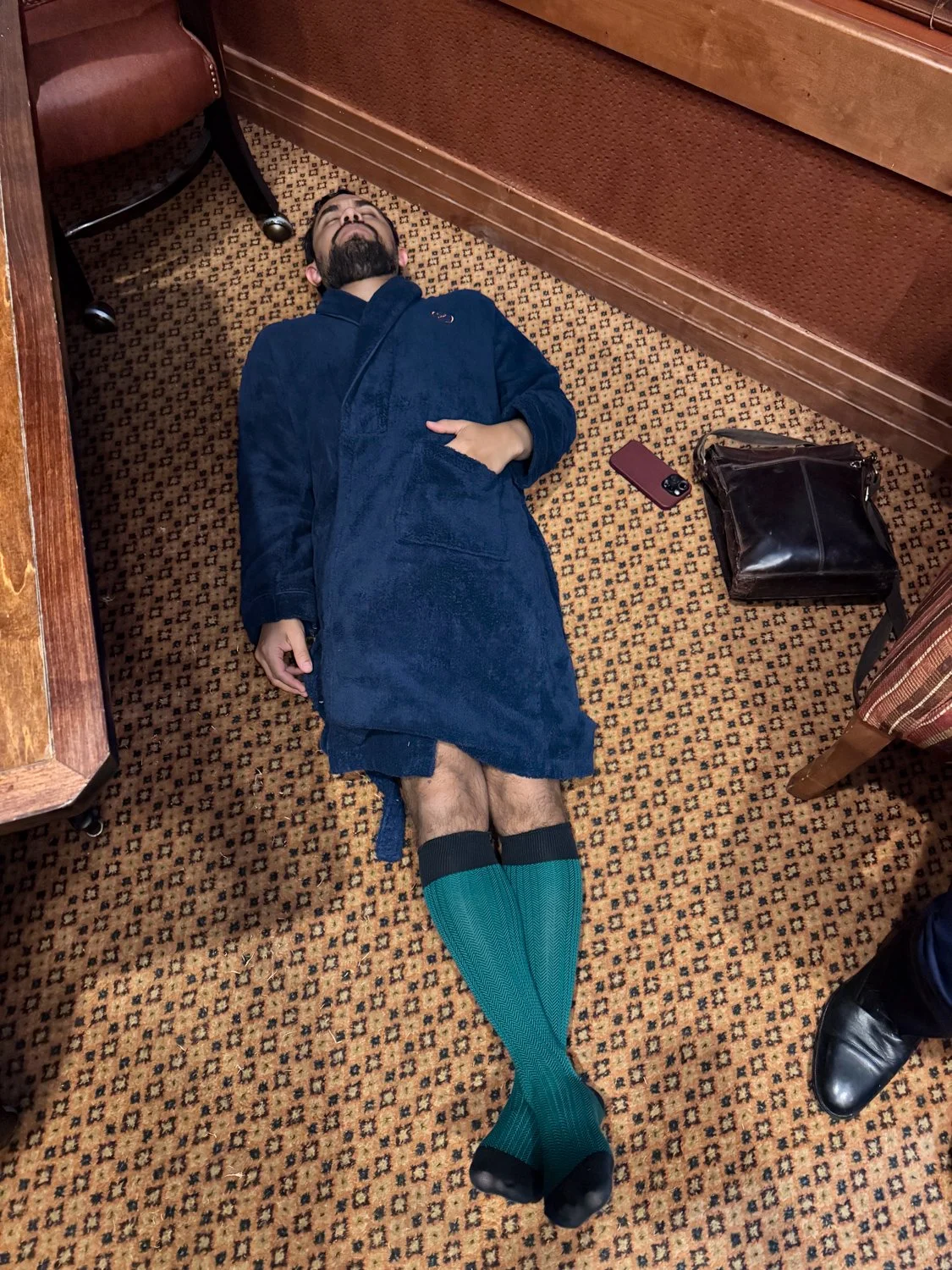 A man lying on the patterned carpet of a hotel room floor, wearing a blue robe and teal knee-high socks, with a black bag, a phone, and a wallet nearby.