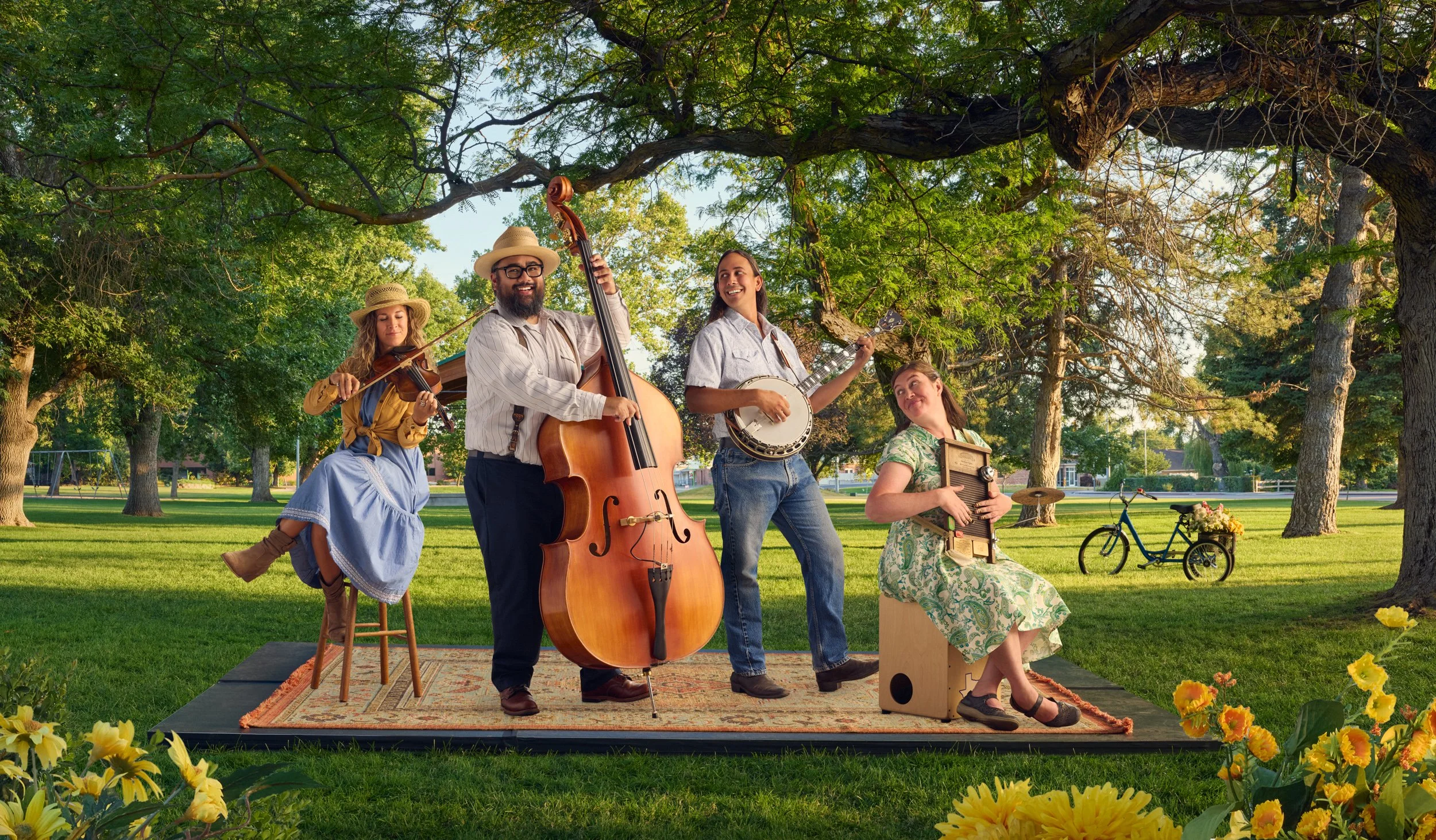 A group of four musicians playing instruments outdoors under a large tree in a park during daytime. There is a girl playing the violin, a man playing the upright bass, a girl playing a banjo, and a woman sitting on a box playing a washboard. The scen